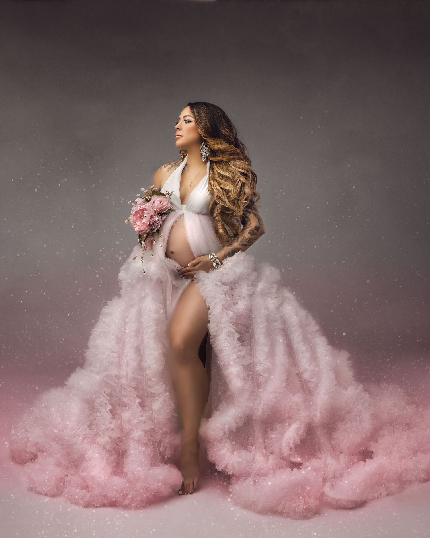 Pregnant woman in flowing pink tulle robe holding flowers during elegant maternity photo shoot.
