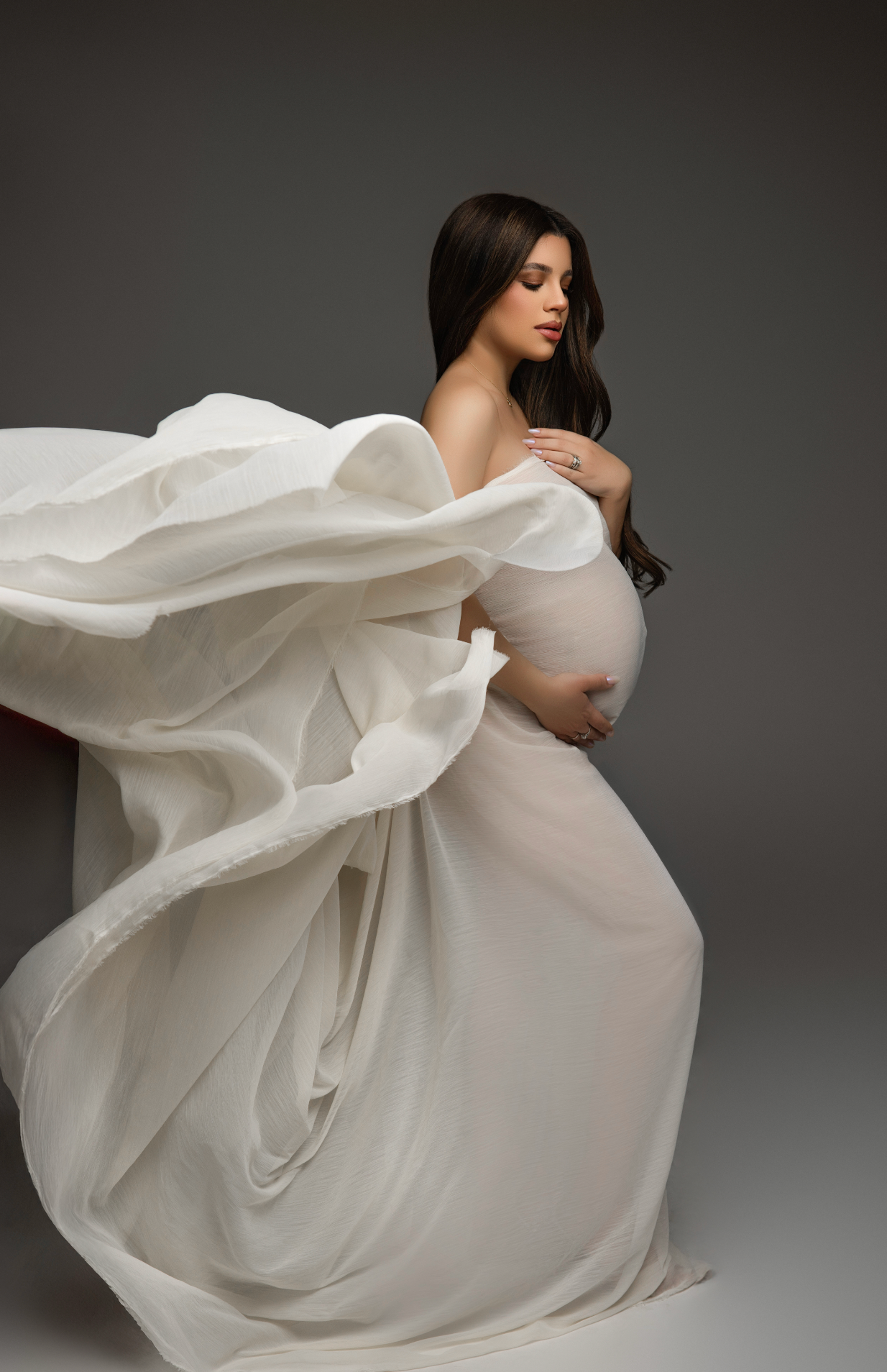 Pregnant woman in flowing white chiffon fabric gown posing gracefully in a studio maternity photography session.