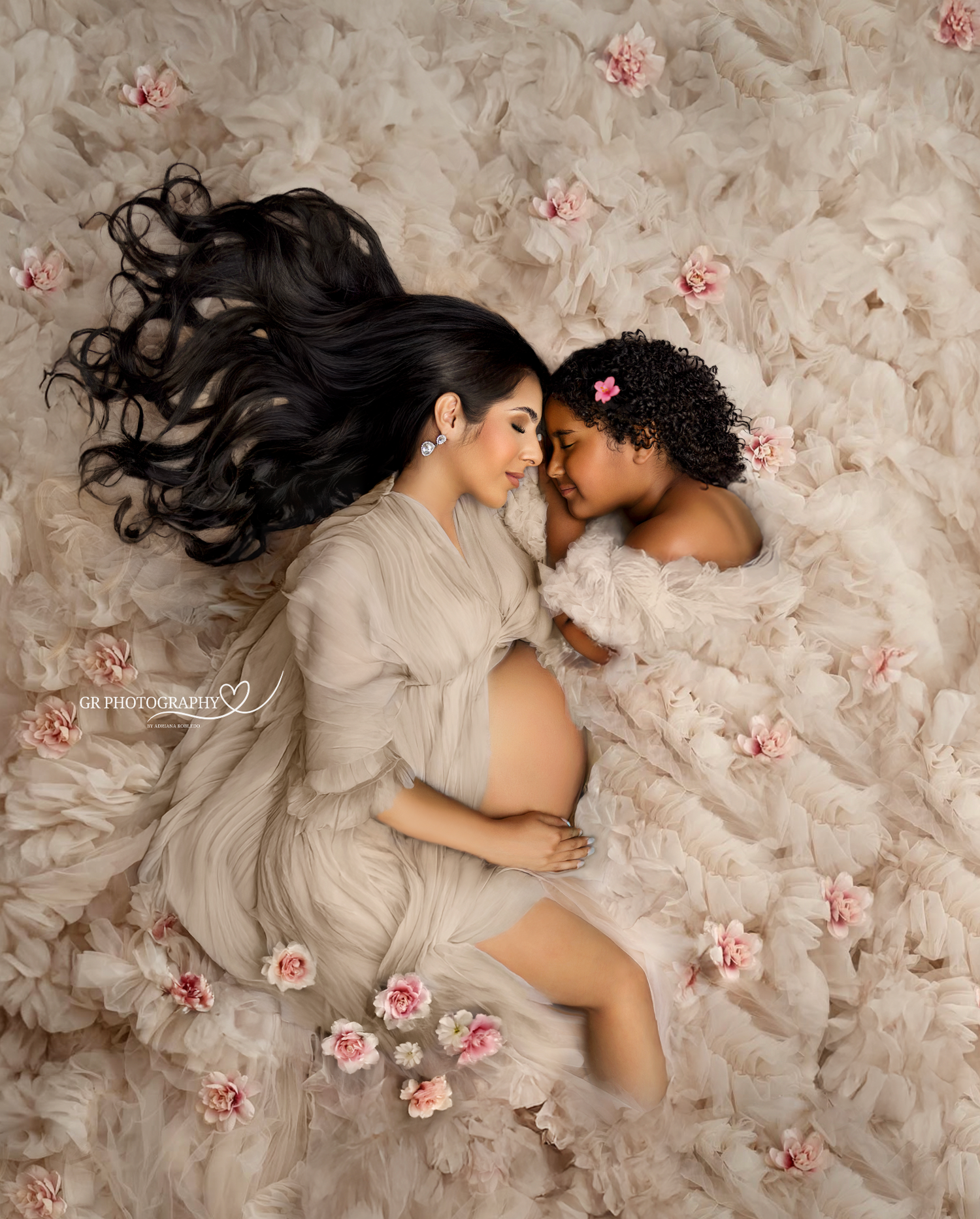 Pregnant woman and young girl lying on floral backdrop in matching beige robes during maternity photo shoot.