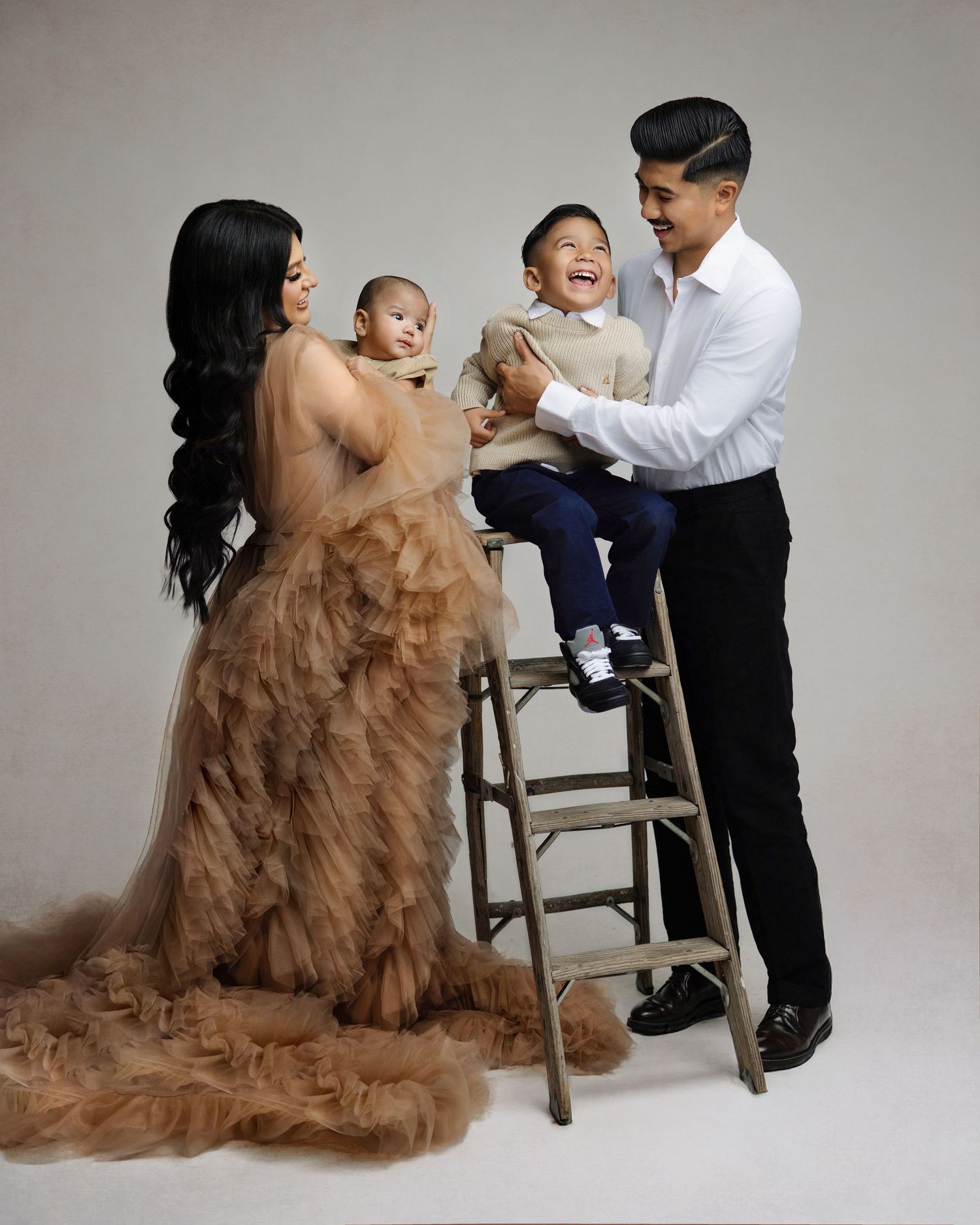Family maternity photo shoot with mother in tulle gown holding baby, father in white shirt with toddler on ladder.
