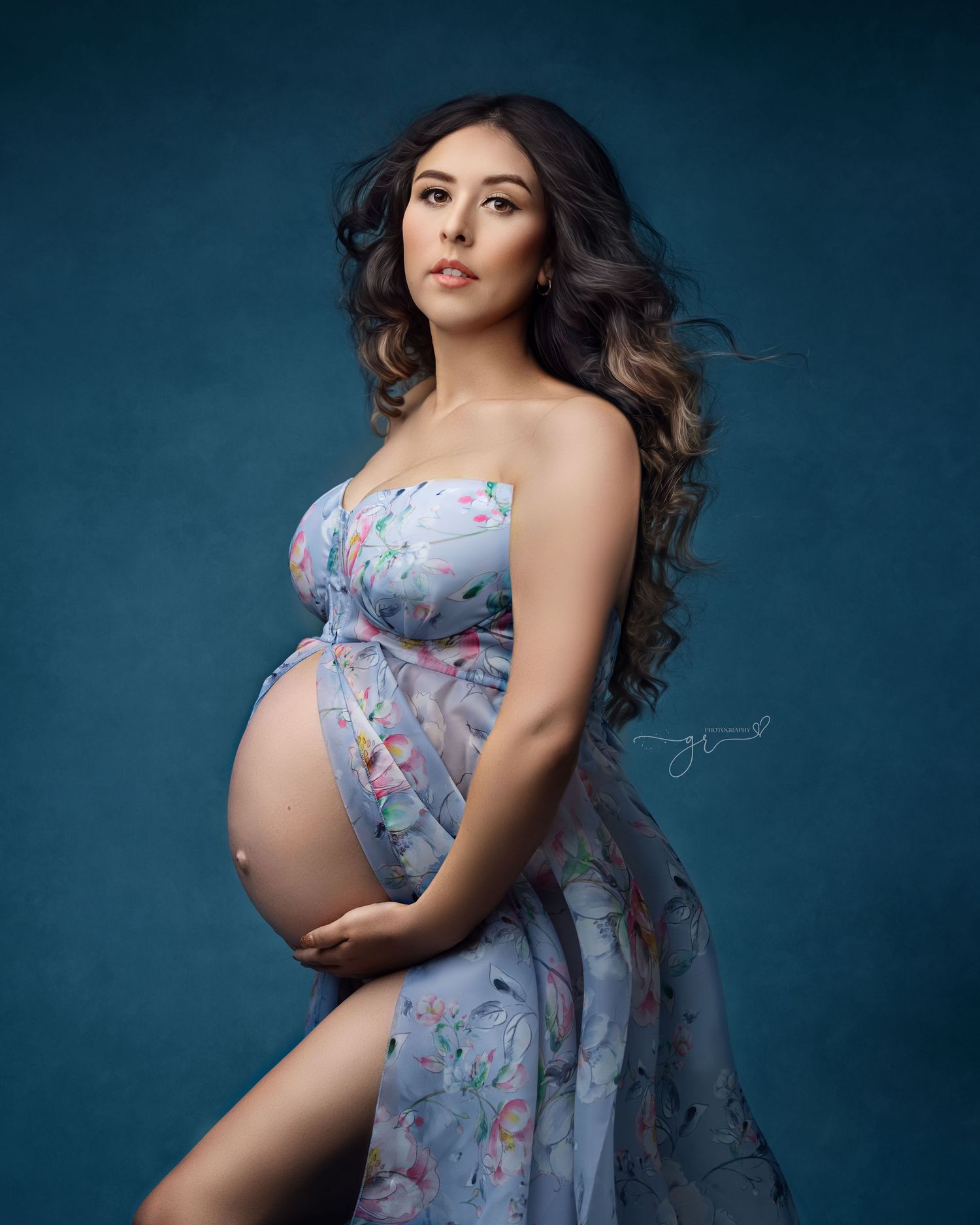 Pregnant woman in floral blue maternity gown cradling baby bump against teal studio background.