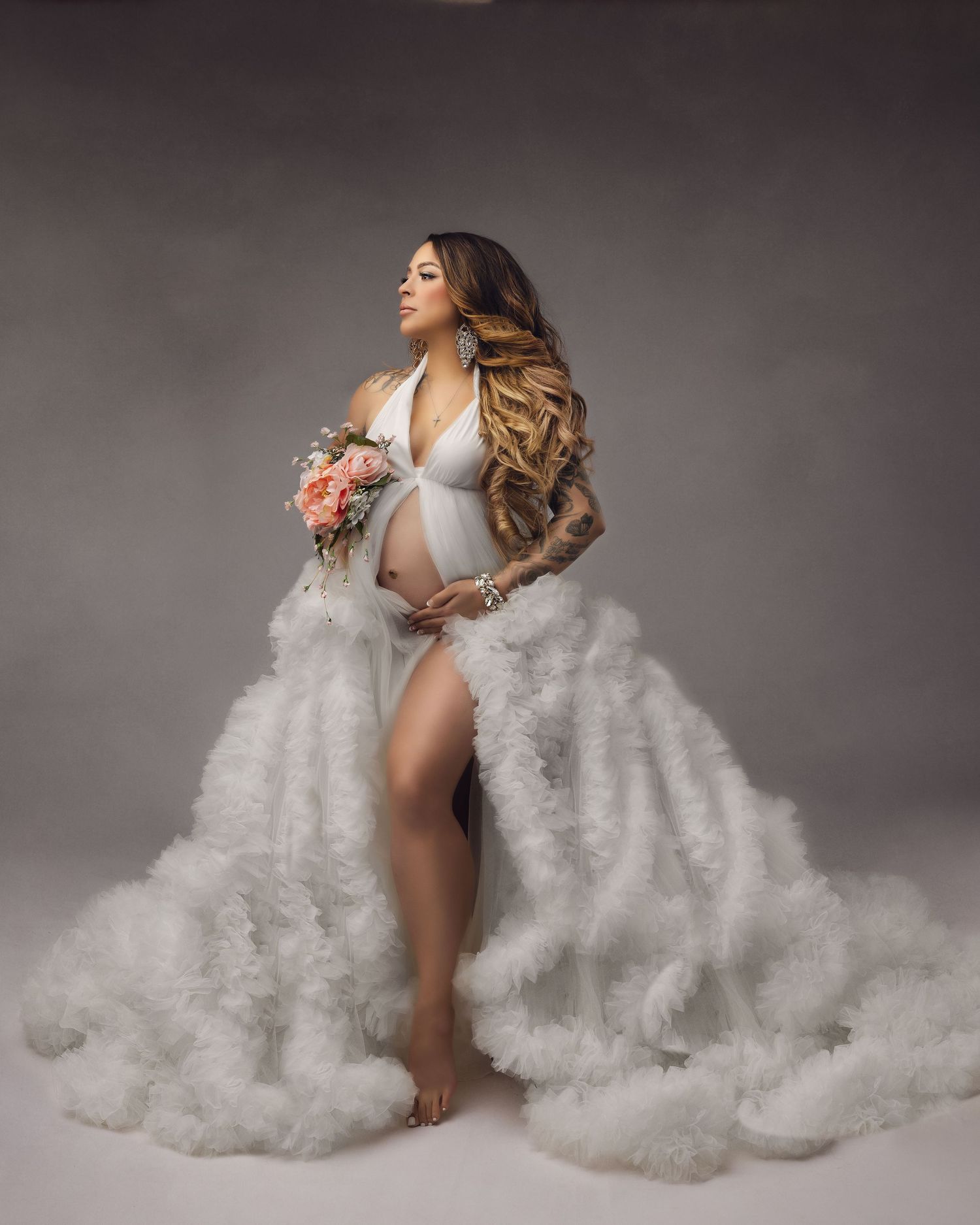 Pregnant woman in white ruffled maternity gown holding floral bouquet, elegant studio maternity photography session.