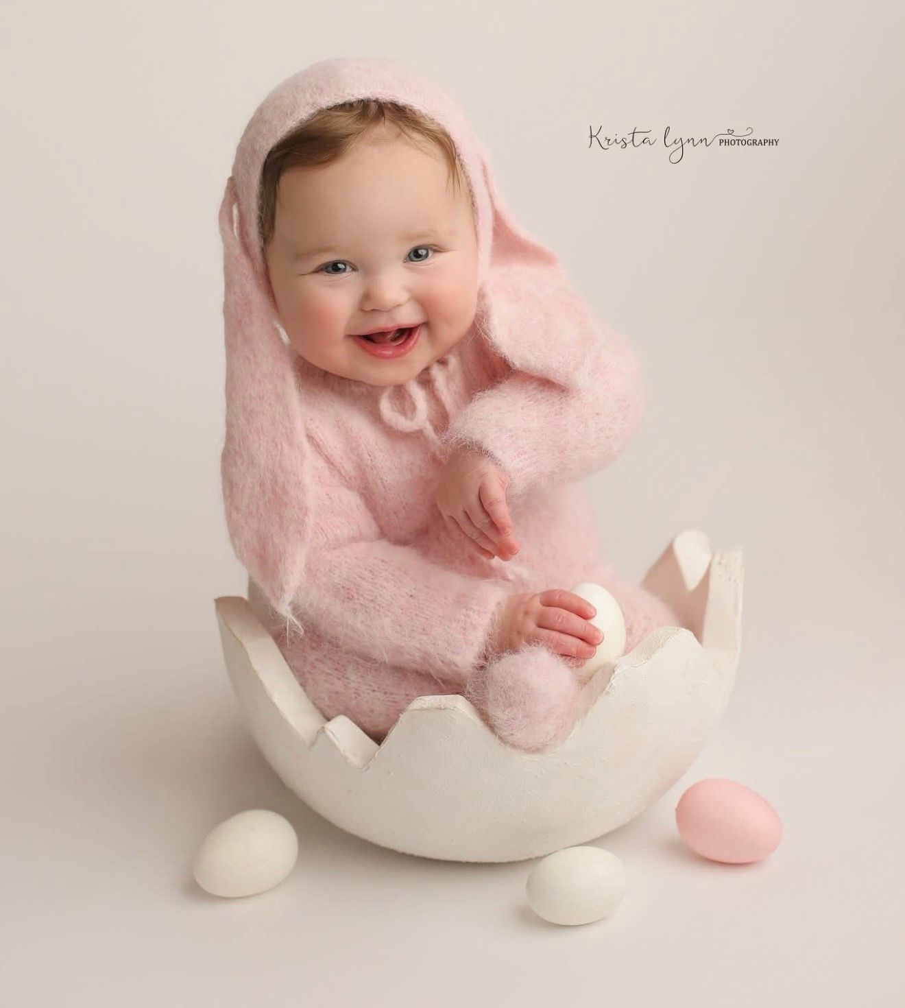 Baby in soft pink outfit sits in white eggshell prop during sweet Easter photo session.