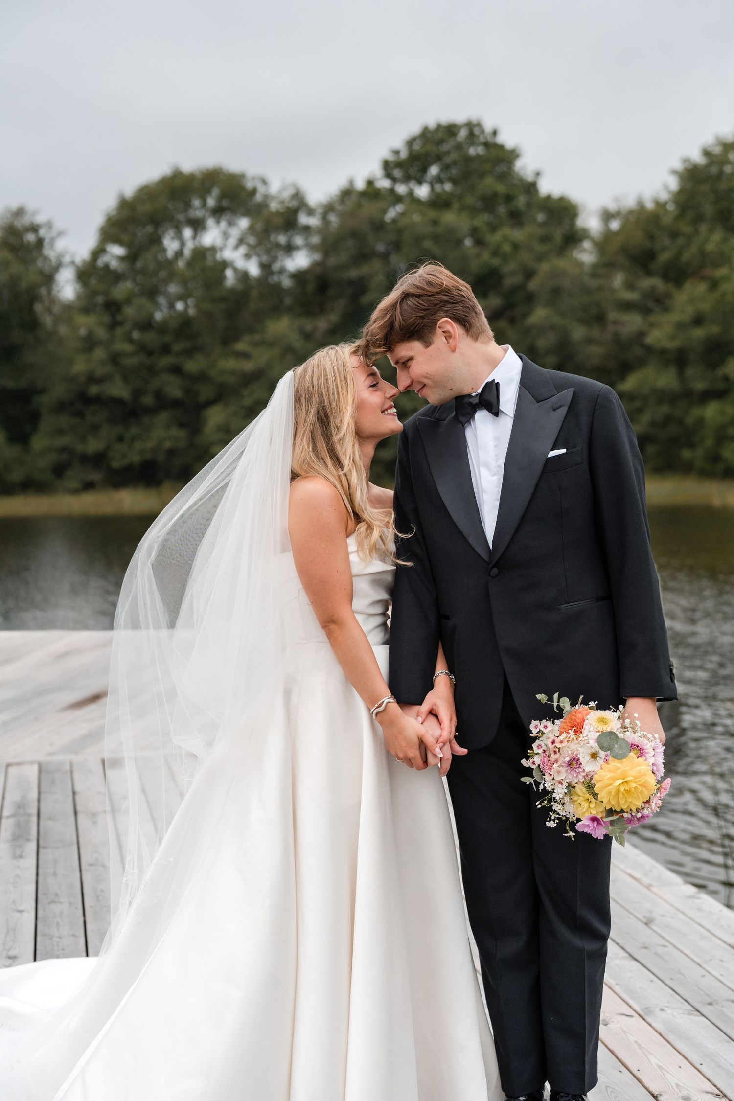 A wedding couple shares tender moments on a wooden dock by a serene lake surrounded by trees.