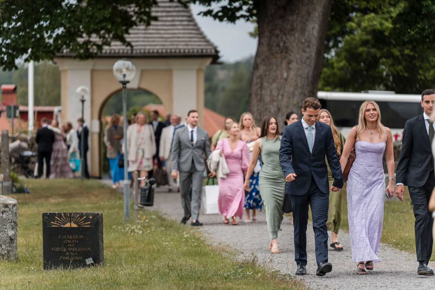 Wedding party walks down a tree-lined path at an outdoor venue during a summer celebration.