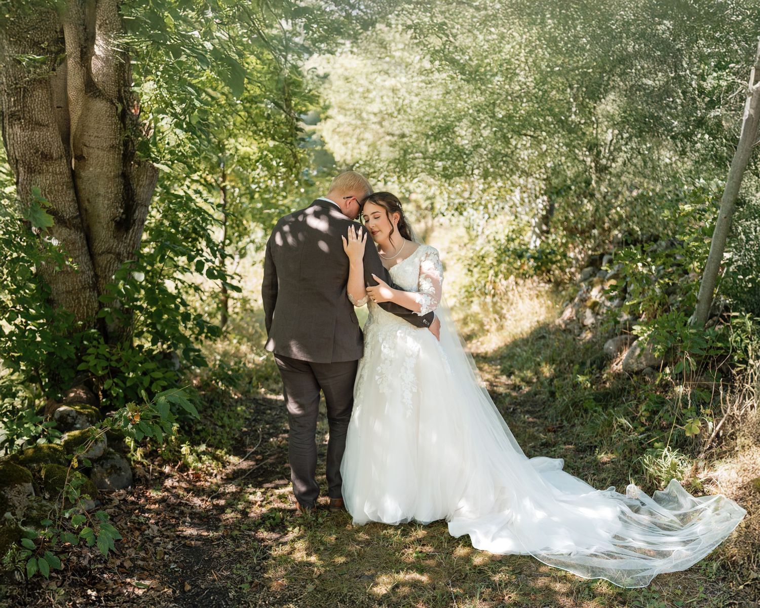 A romantic wedding photoshoot in a sunlit wooded area with a flowing white gown trailing on the forest floor.