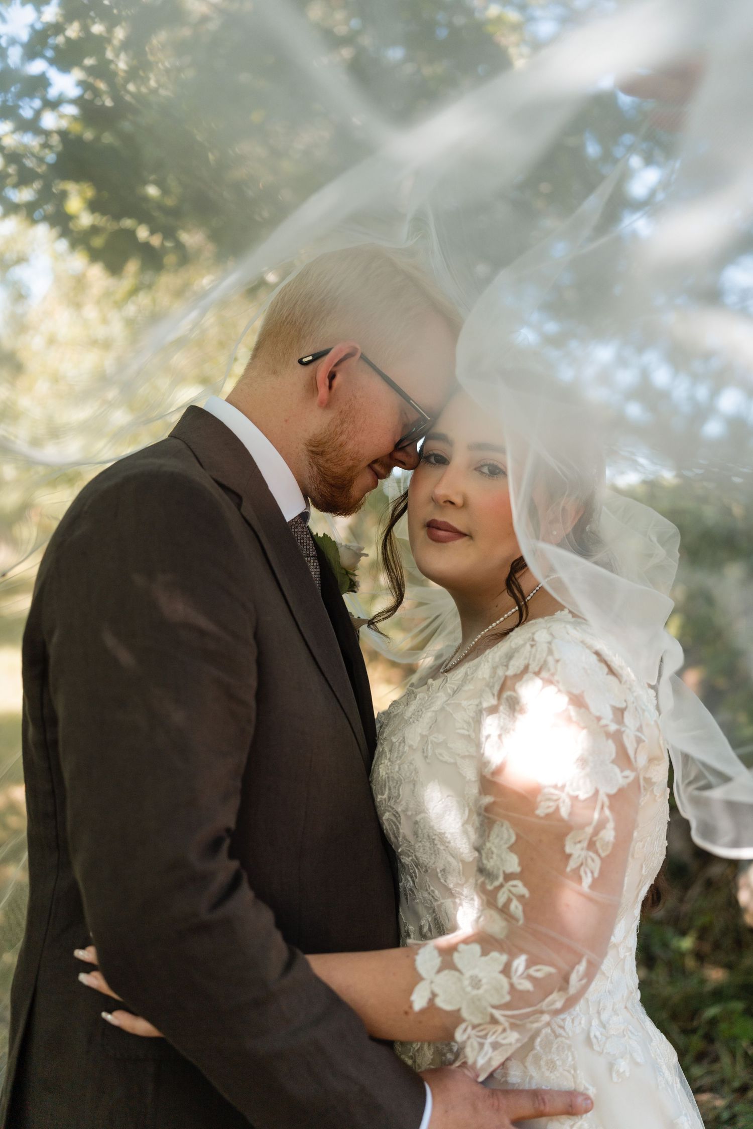 A tender moment between newlyweds under a delicate veil with sunlight filtering through the trees.
