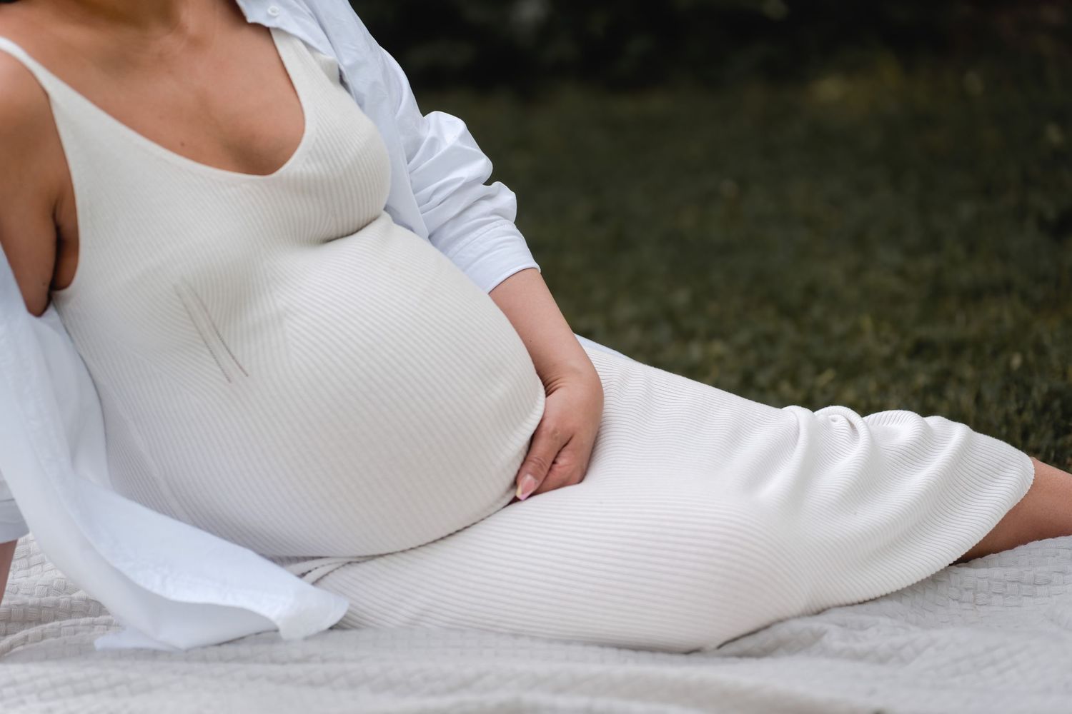 A person wearing a white dress sits on a white blanket outdoors in a relaxed pose.
