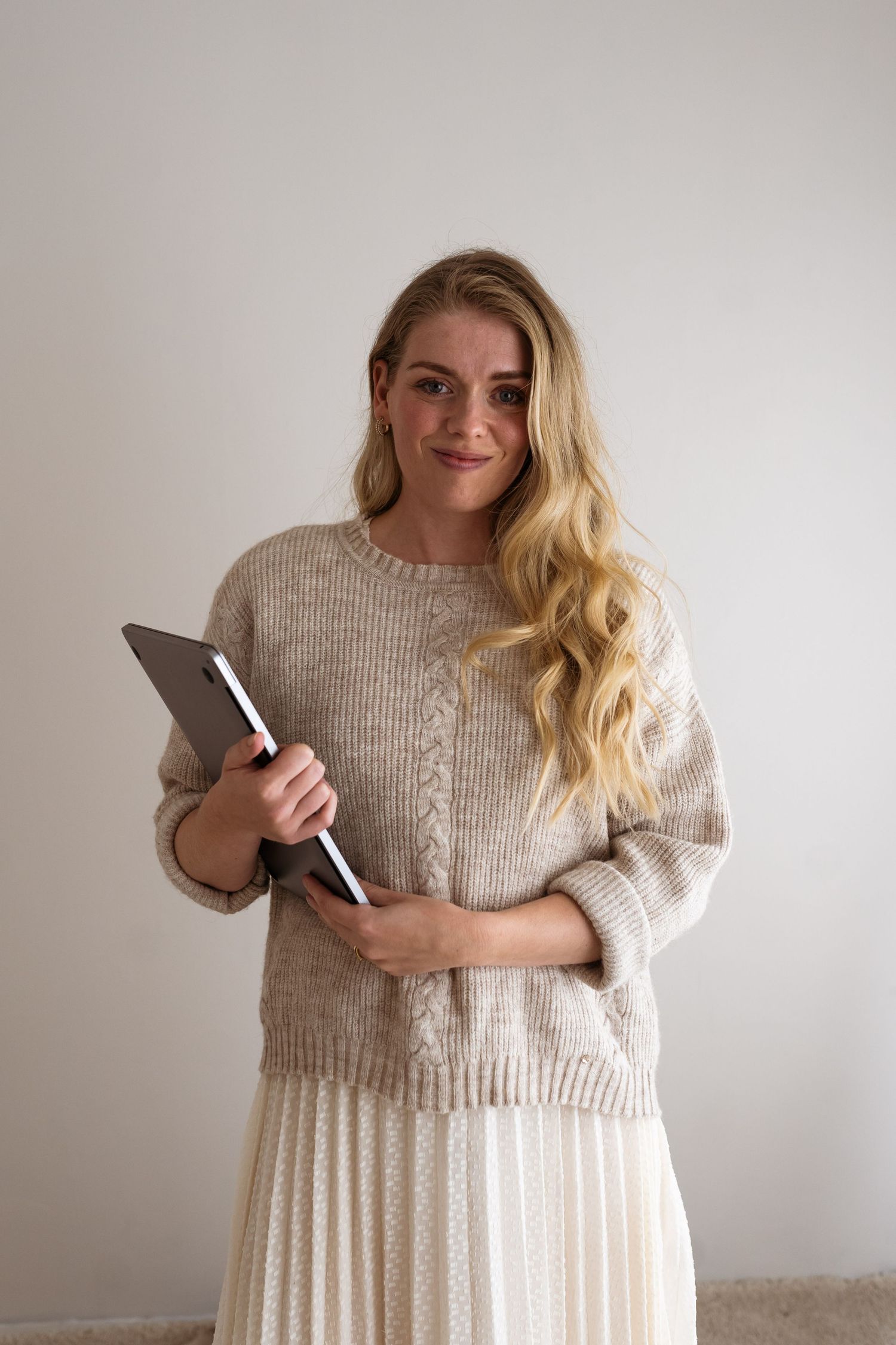 A person in a cozy beige sweater and pleated white skirt holds a tablet while standing against a neutral wall.