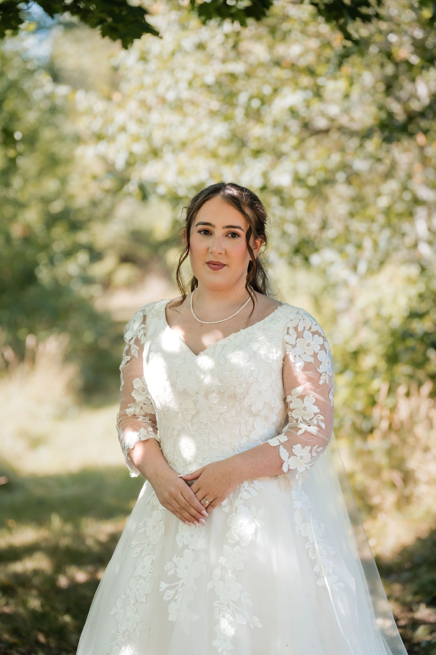 A bride in a white lace wedding dress with sheer sleeves poses outdoors among blooming spring trees.