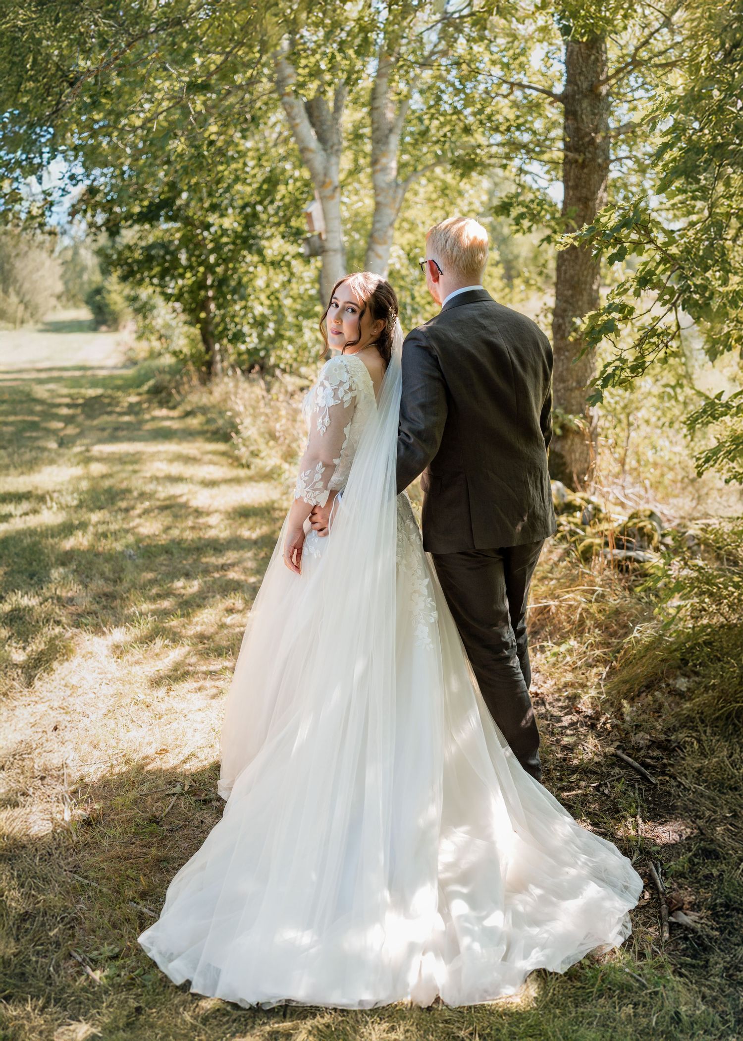 A bride and groom share intimate moments in a sun-dappled outdoor setting with trees in the background.