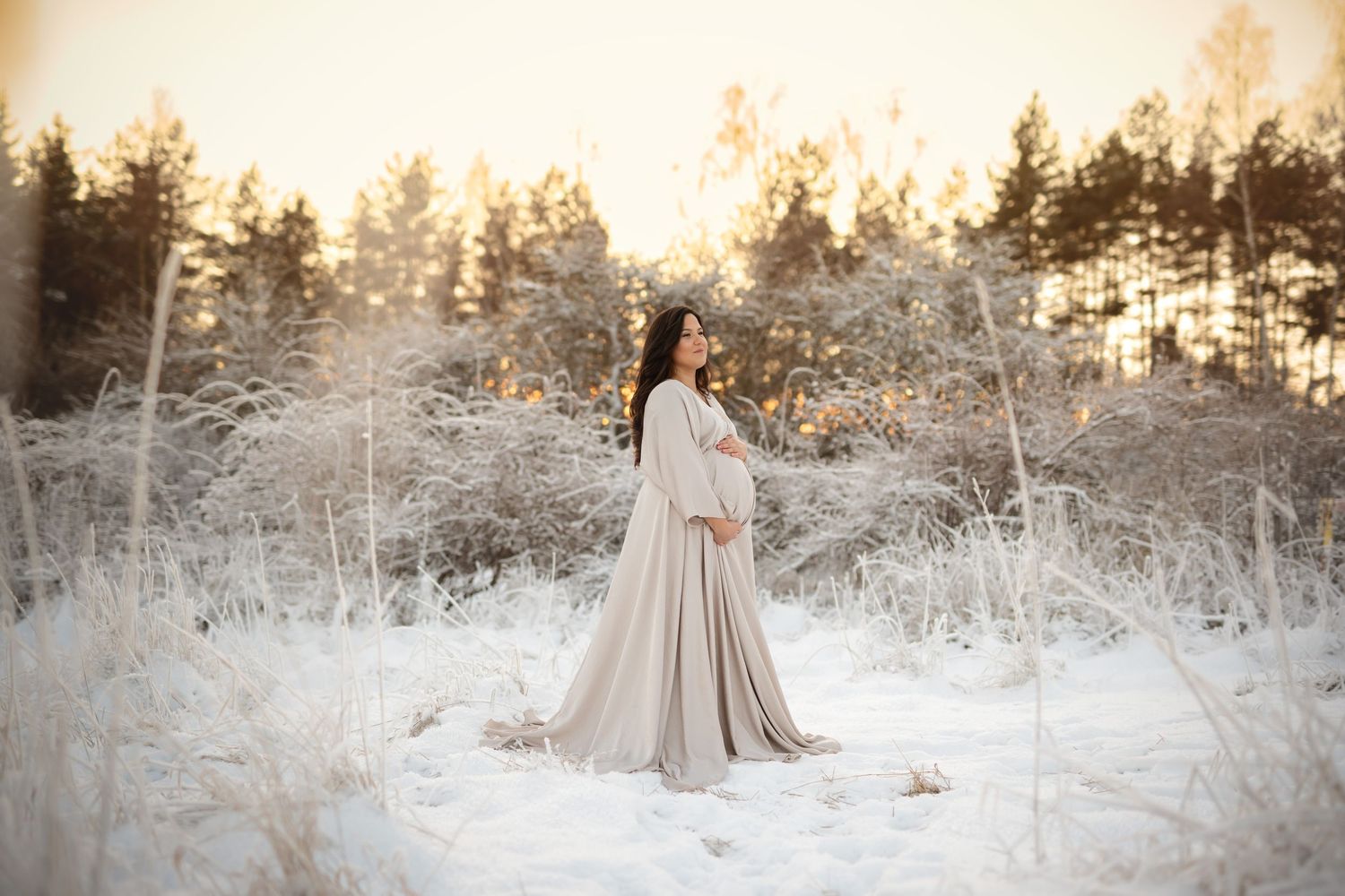 A figure in a flowing beige dress stands in a snowy winter forest landscape at sunset.