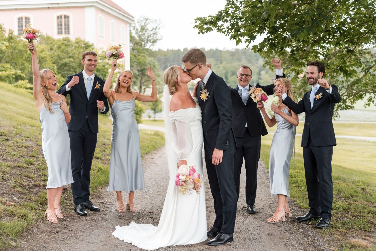 Wedding party in pastel dresses and navy suits poses together outdoors on a sunny summer day.