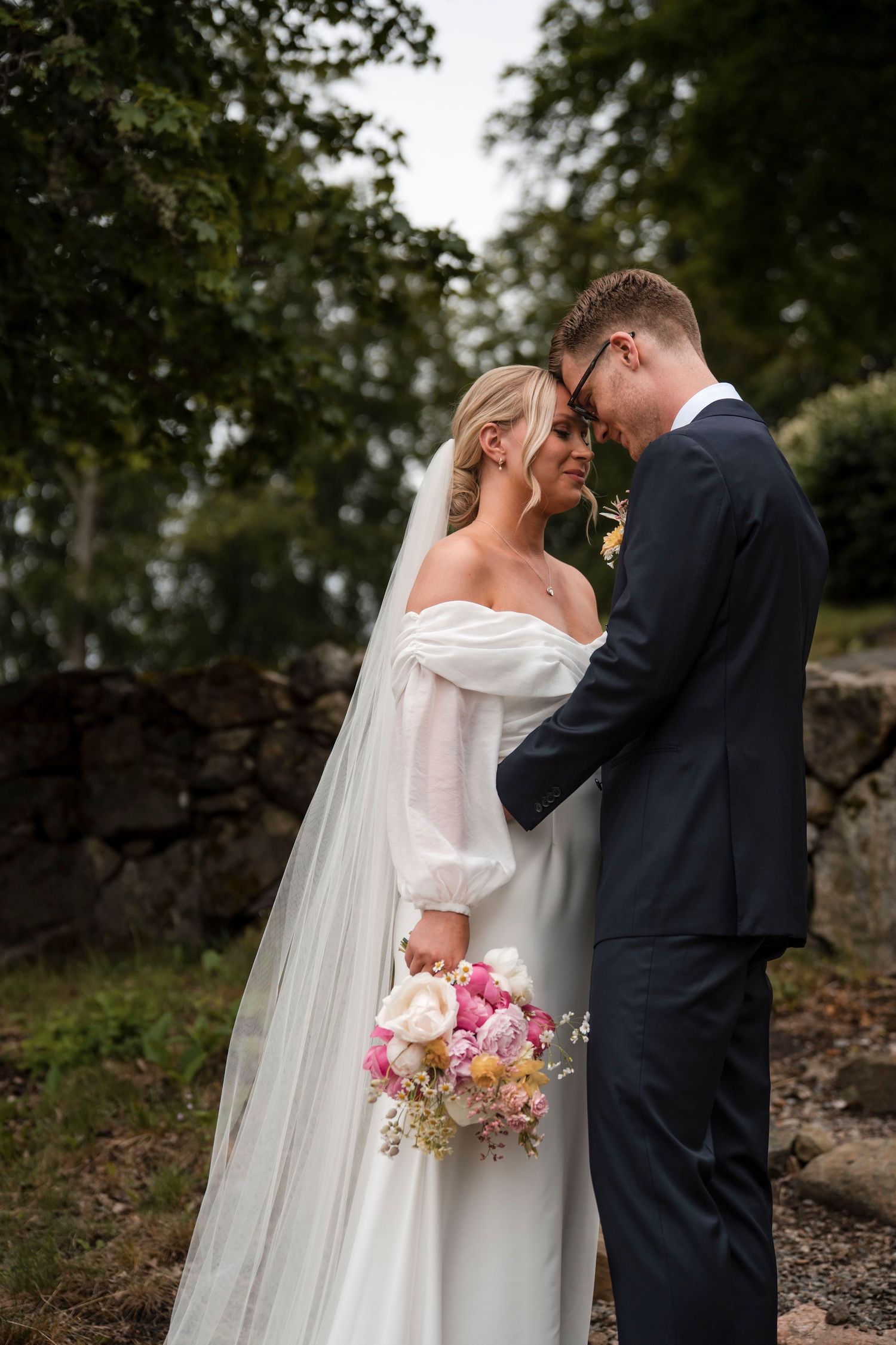Bride and groom share an intimate moment outdoors with pink bouquet against a rustic stone wall backdrop.