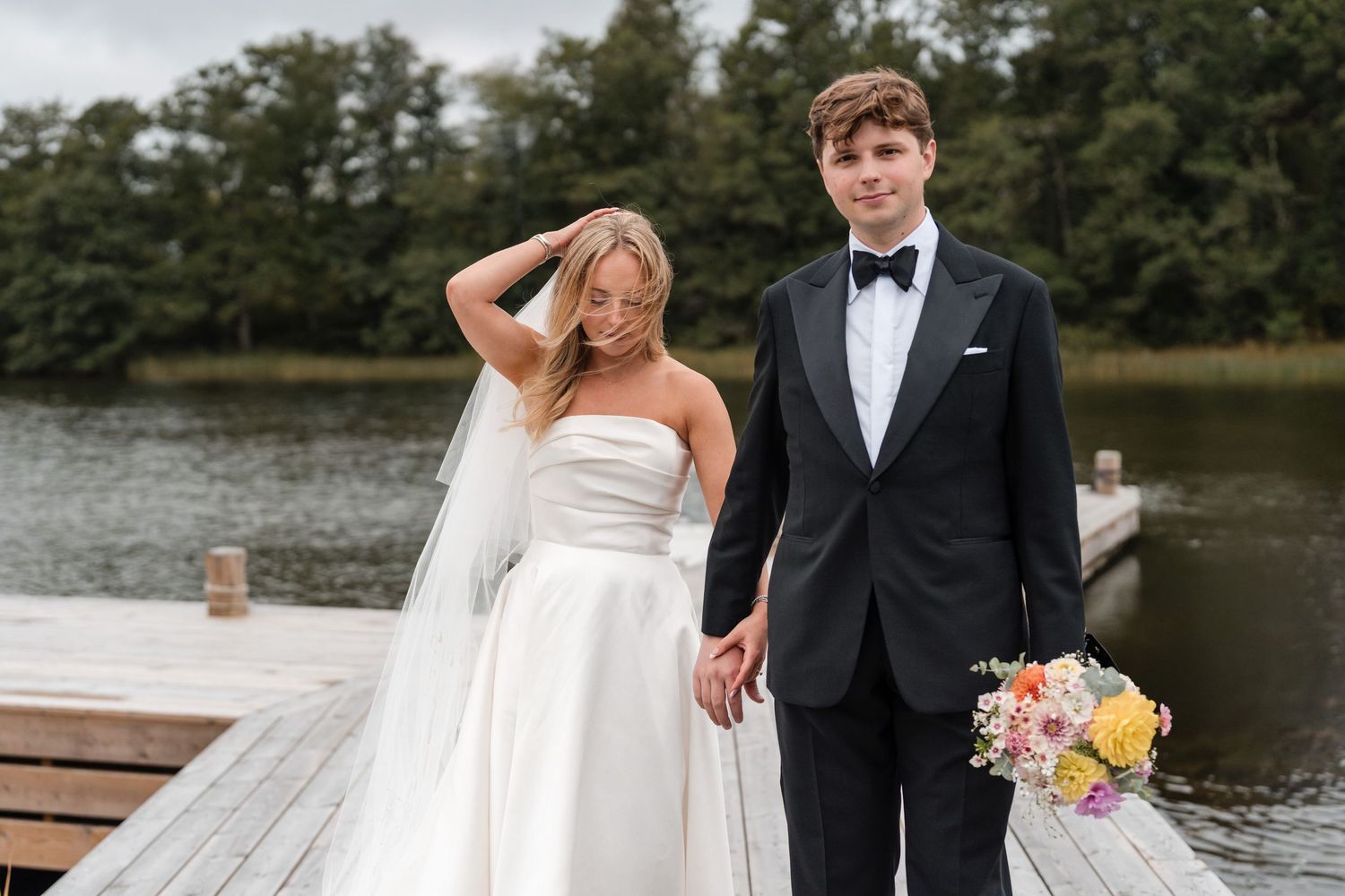 A bride and groom share romantic moments by a lake on their wedding day in formal attire.