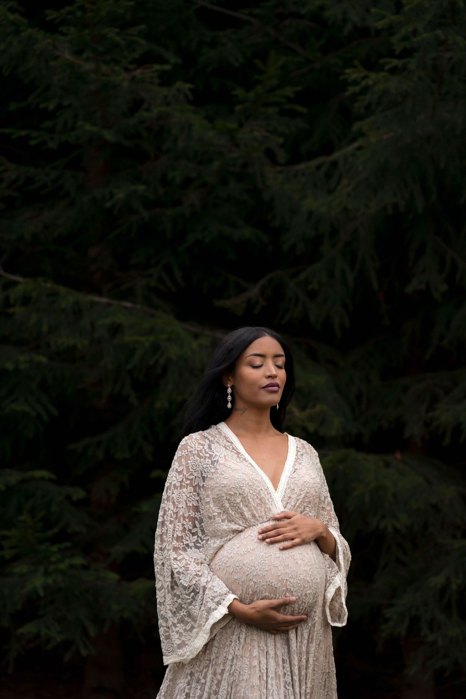 A woman in a white lace dress poses elegantly against a dark forest backdrop in a maternity photoshoot.