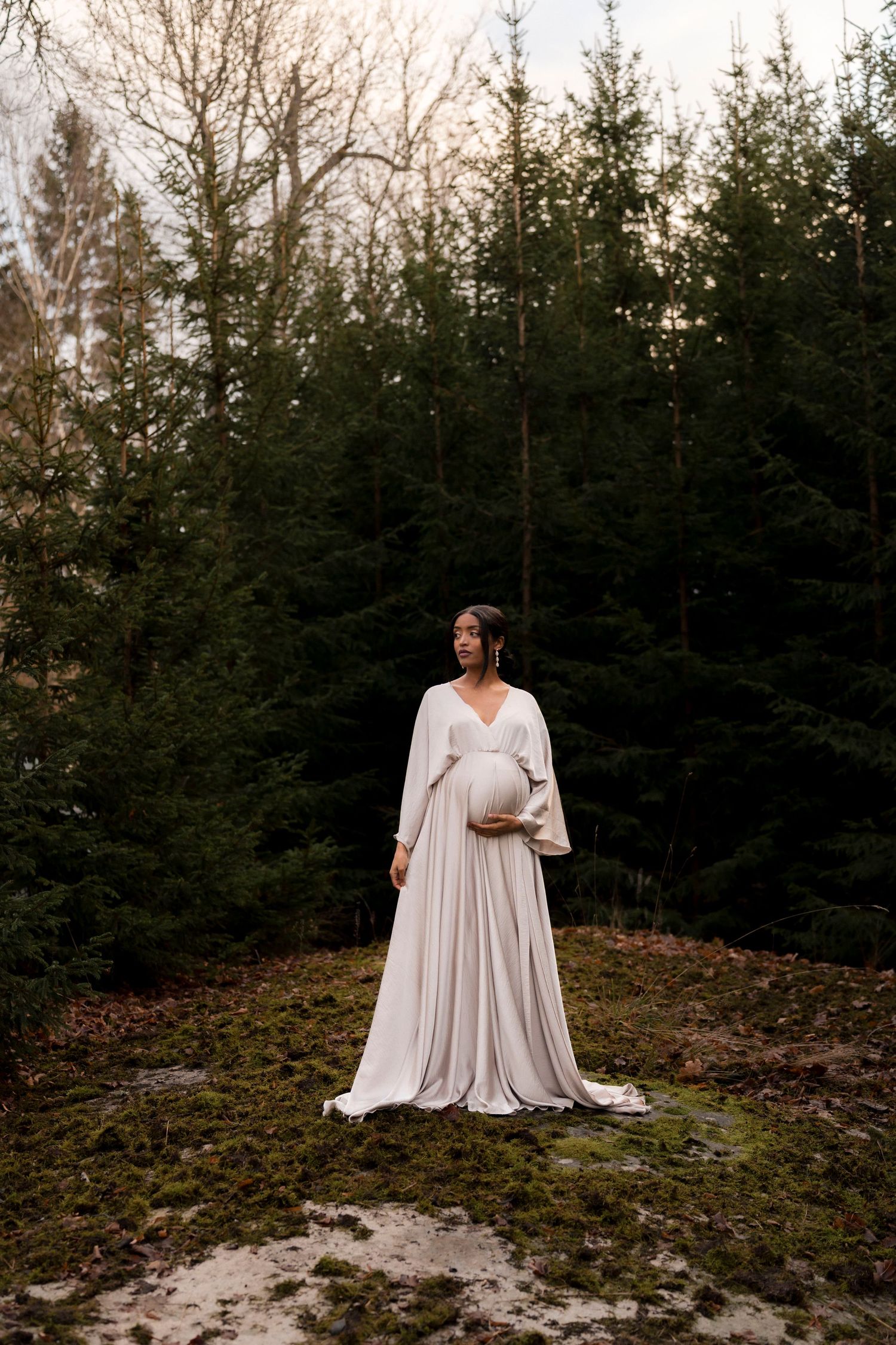 A figure in a flowing white dress poses gracefully against a backdrop of evergreen trees in a moody outdoor setting.