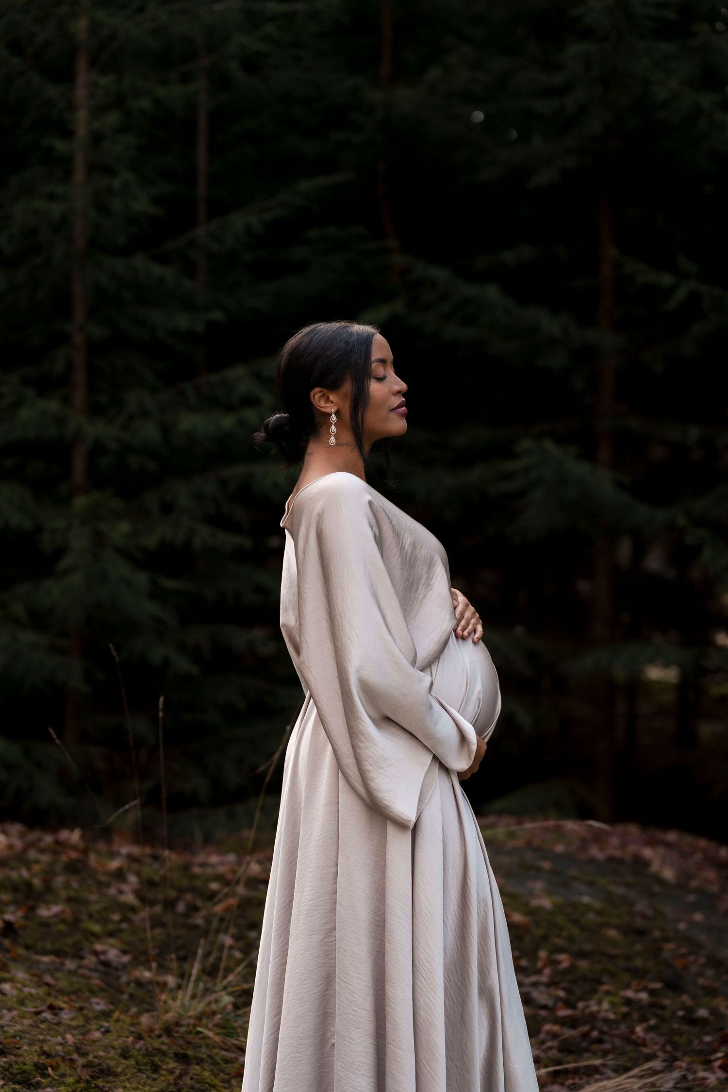 A person in a flowing white dress stands in profile against a dark forest background, cradling their pregnant belly.