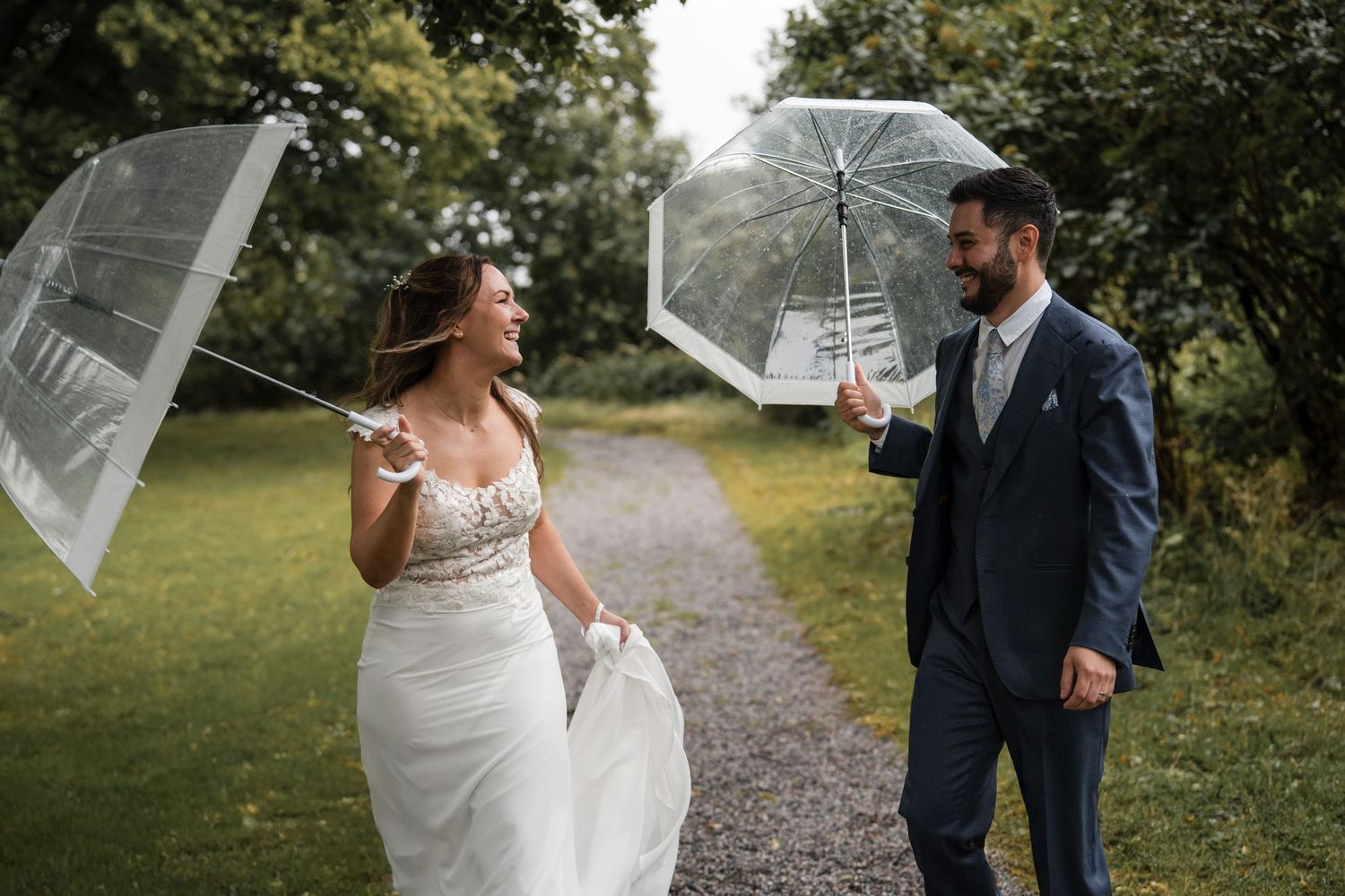 Wedding couple shares romantic moment on garden path while holding clear umbrellas on rainy day.
