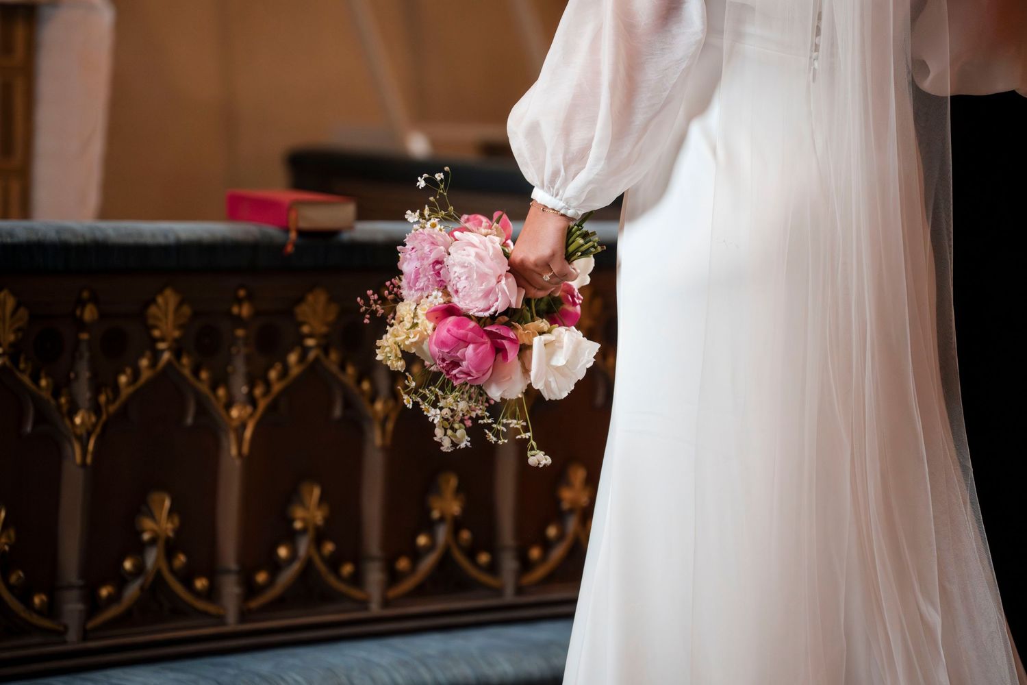 A bride in a white wedding dress holds a pink and white floral bouquet against an ornate wooden railing.