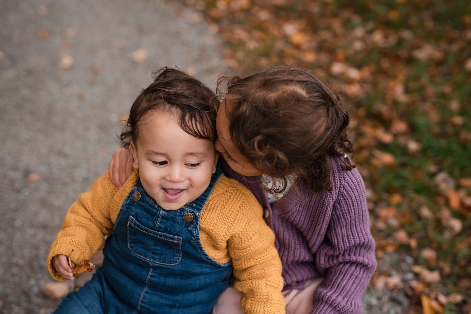 Two children share a sweet moment together on a path surrounded by autumn leaves.