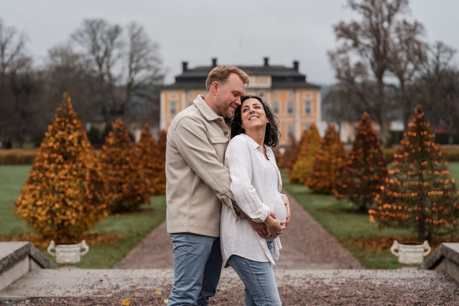 A couple embraces on a path in front of an elegant mansion with autumn trees in the background.