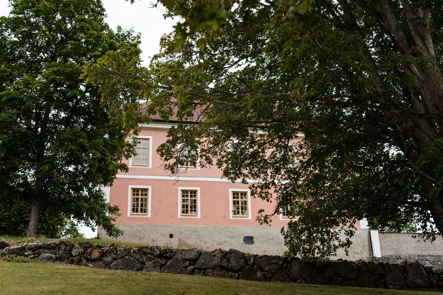 A pink ranch-style house with white trim nestled among mature trees and landscaping in a residential setting.