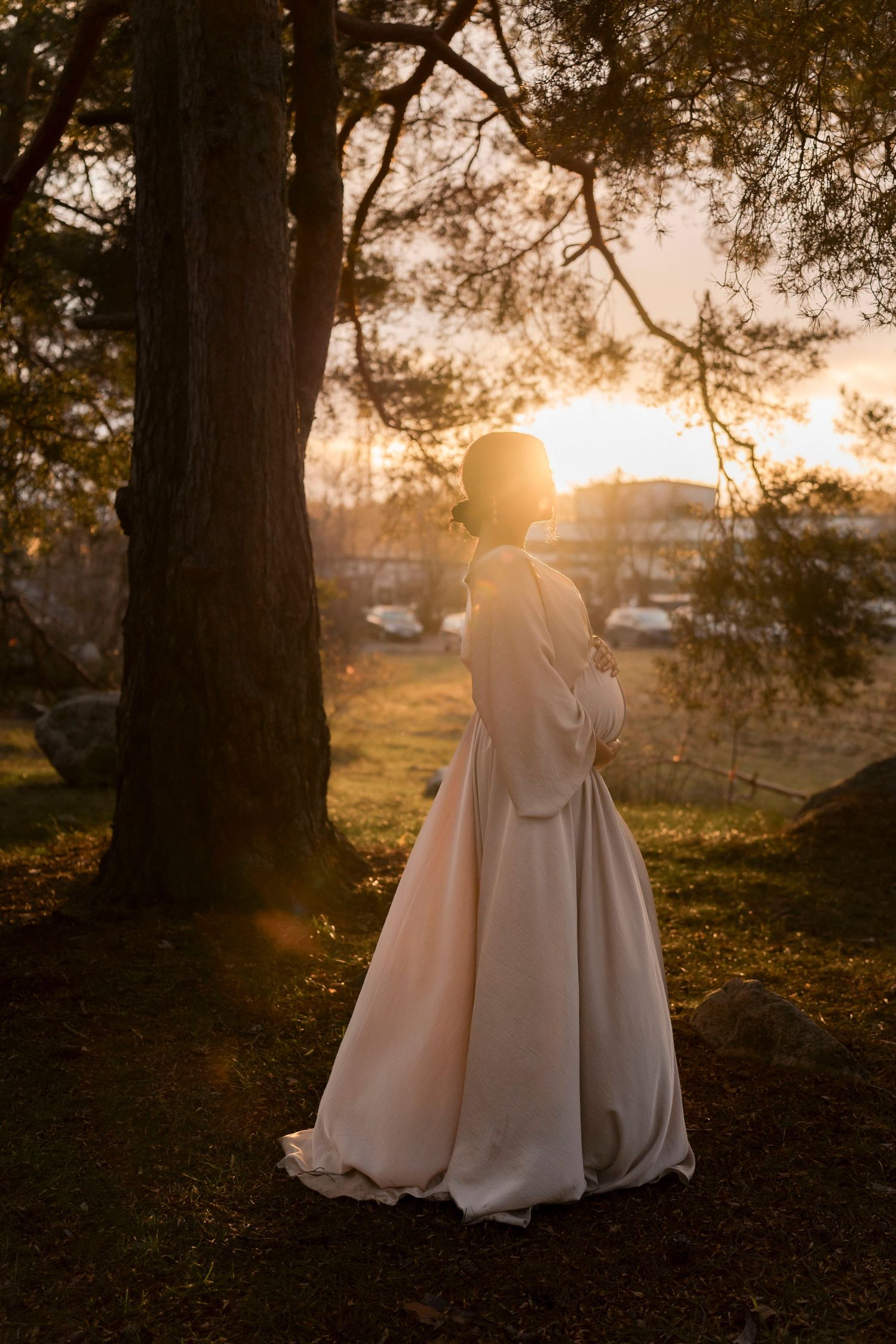 A couple in wedding attire stands silhouetted against a golden sunset beneath a large tree in a romantic outdoor setting.