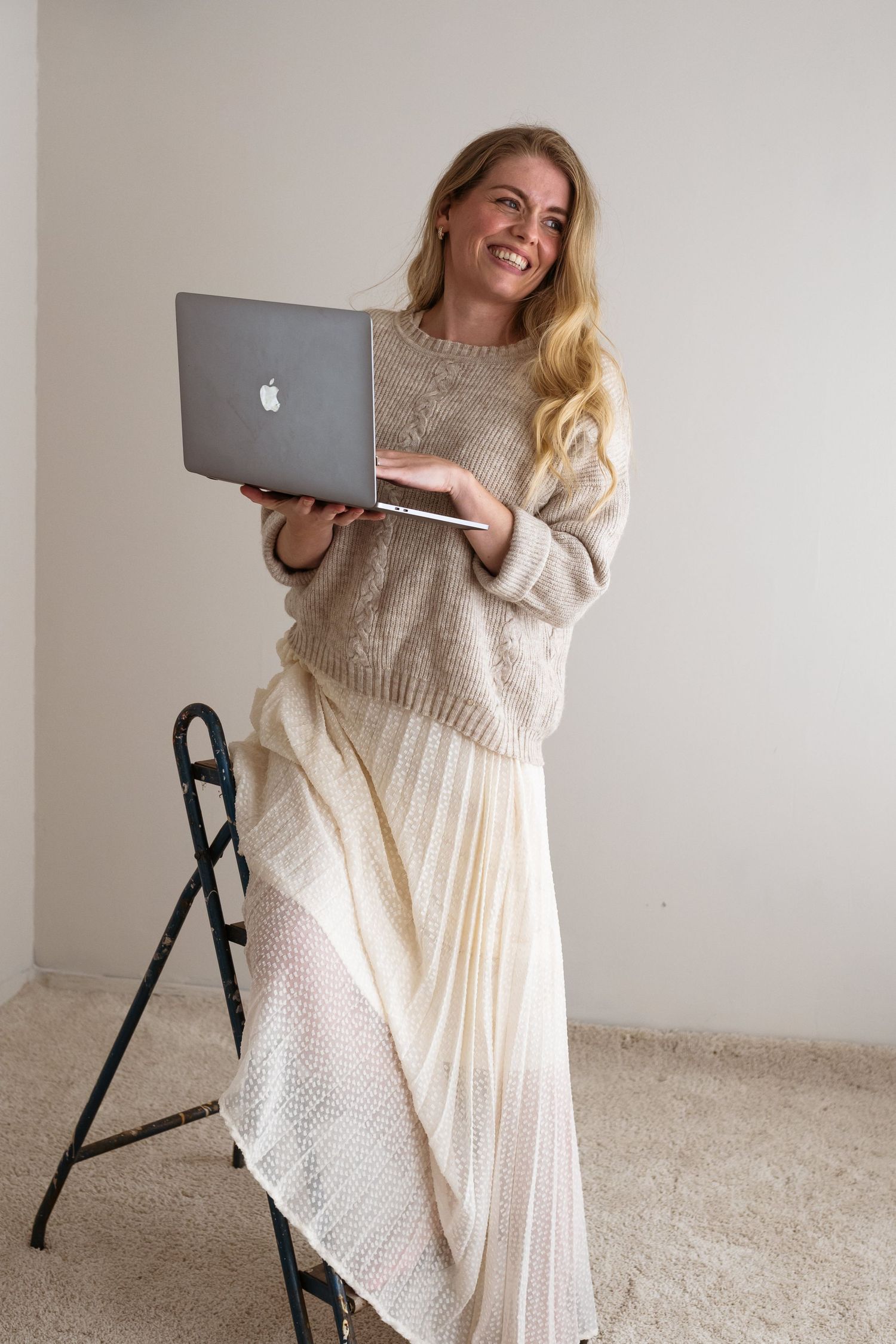 Woman in cozy beige sweater and flowy white skirt holds MacBook laptop while smiling in minimal interior setting.
