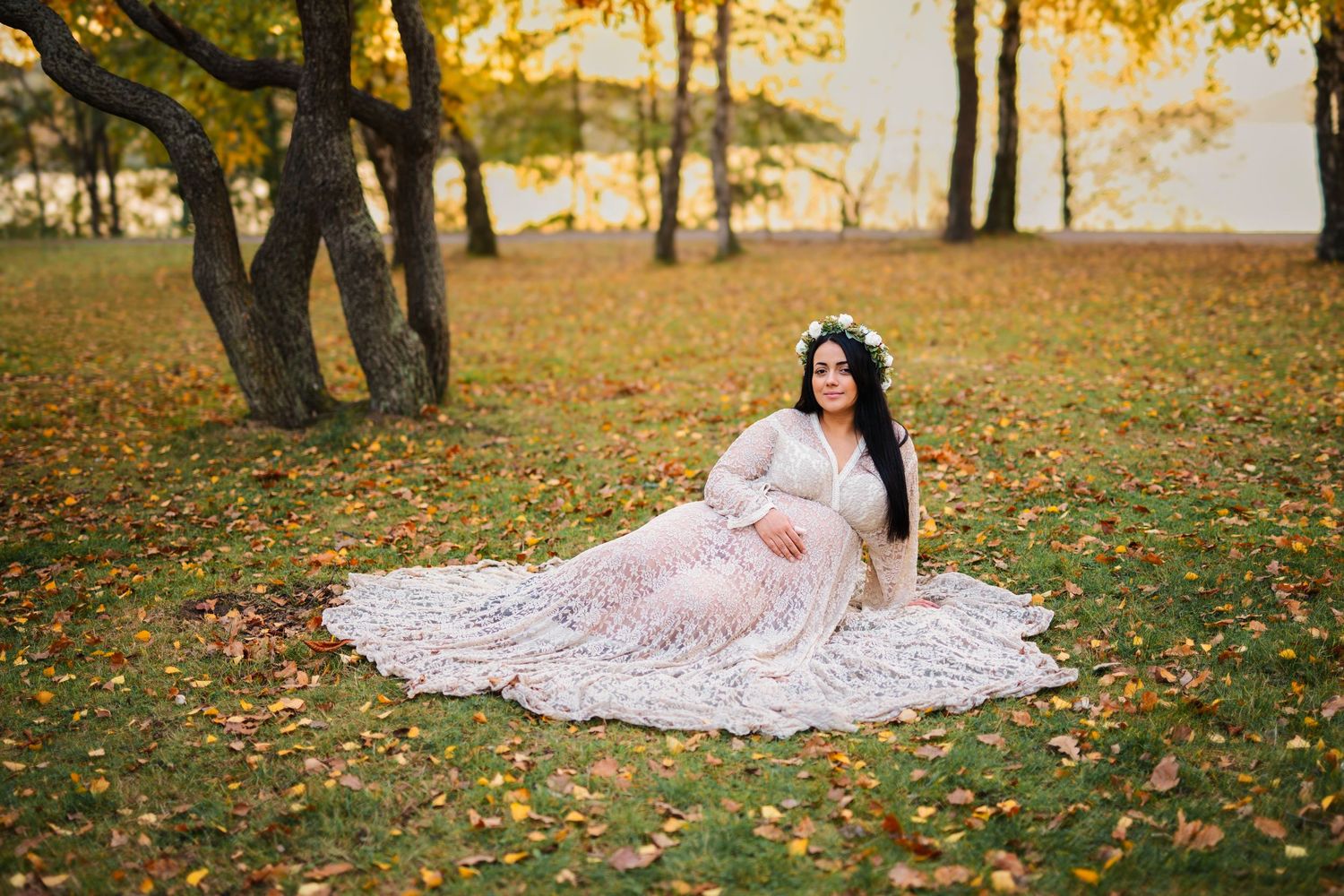 A figure in a white lace gown poses gracefully among autumn leaves in a golden-lit forest setting.