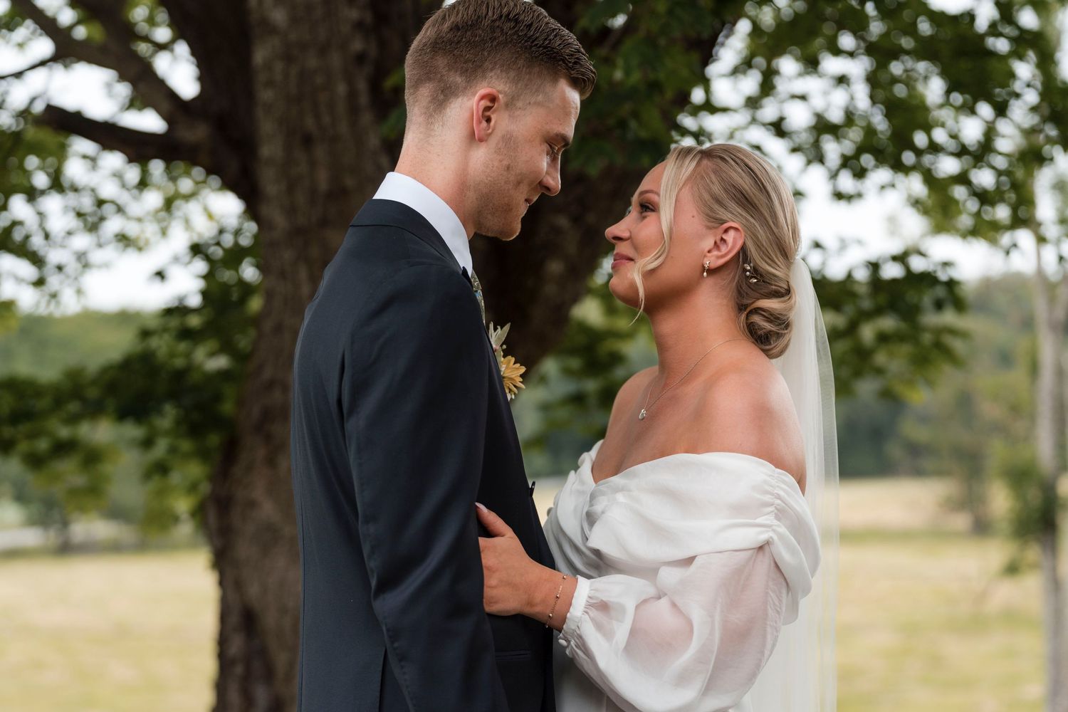 A couple shares intimate moments during their outdoor wedding photoshoot under trees in a natural setting.