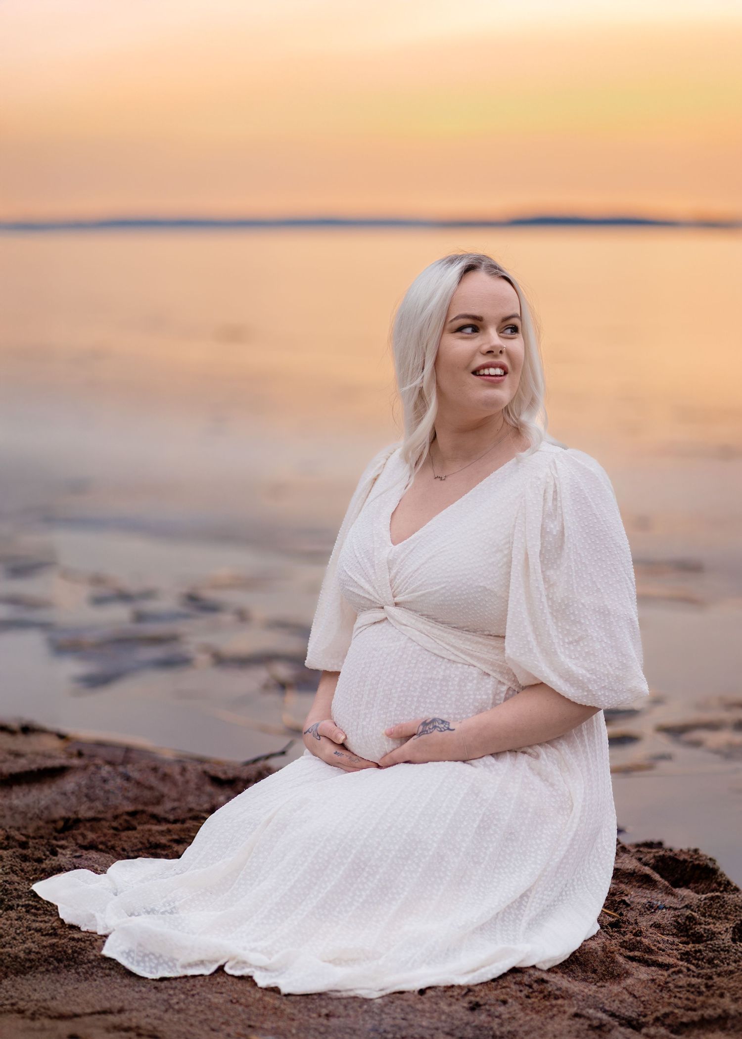 Expectant woman in flowing white dress sits on beach rocks during golden sunset over calm water.
