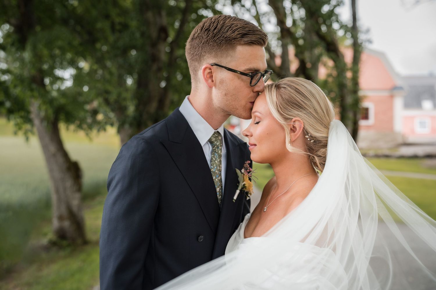 A tender wedding moment captured outdoors with a flowing white veil and elegant formal attire against a natural backdrop.