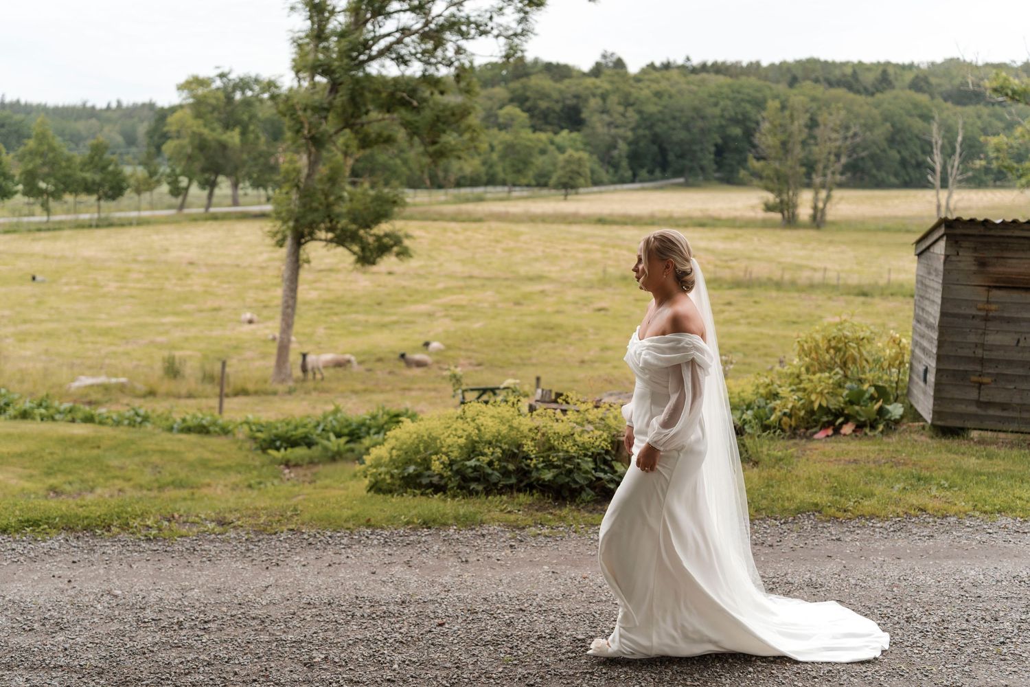 A bride in a flowing white wedding dress walks through a rustic countryside setting during a photo session.