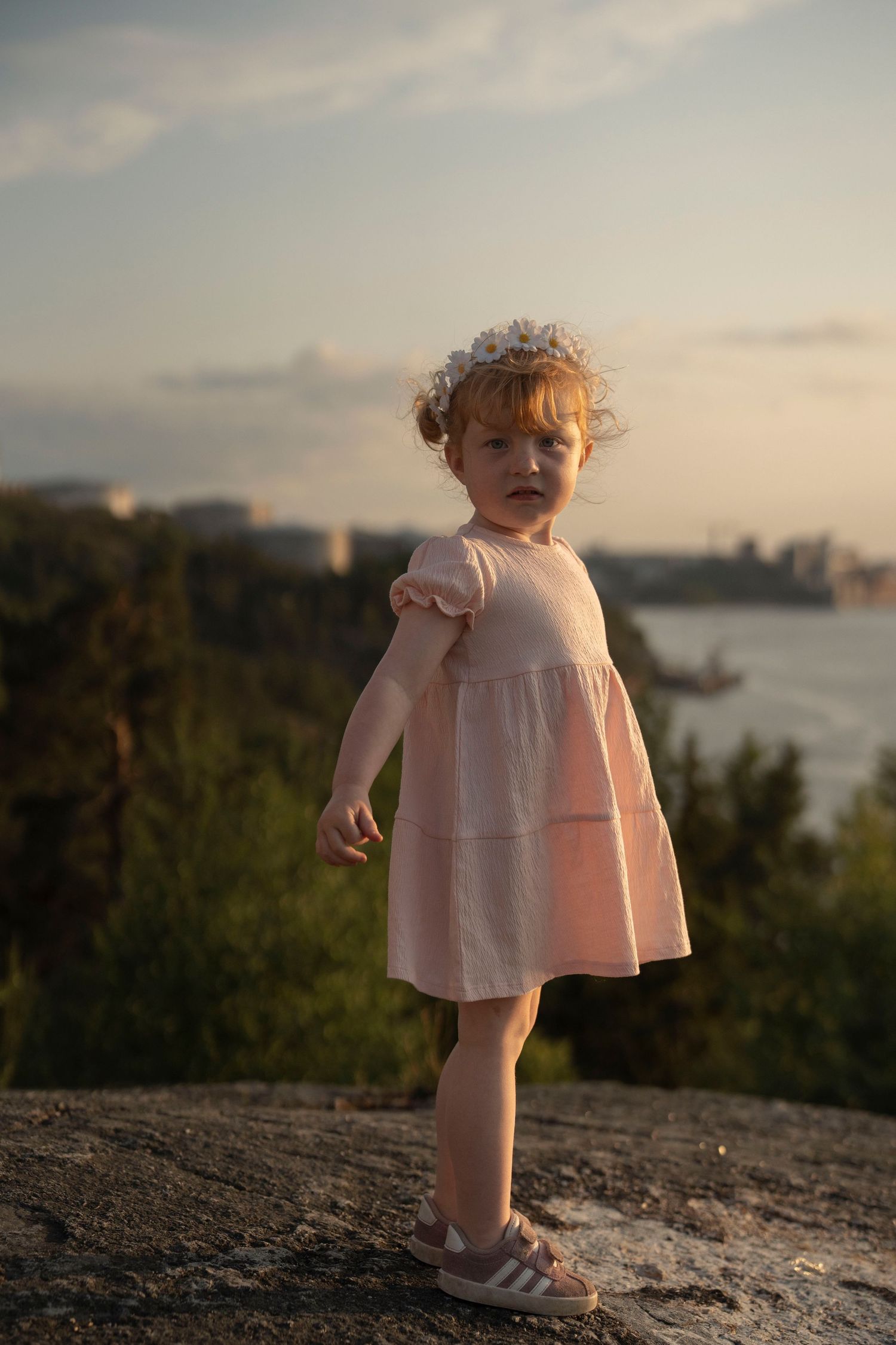 A small child in a pink dress stands on a rocky overlook at sunset with the ocean and cityscape in the background.