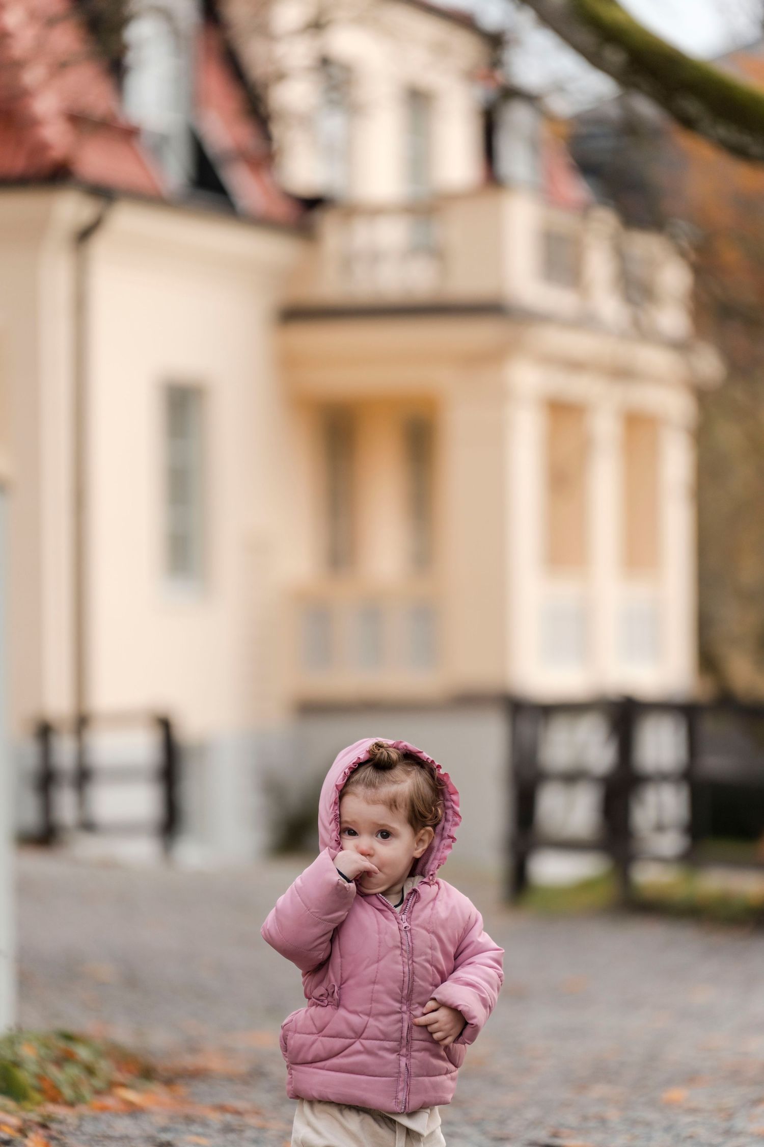 A small child in a pink winter coat plays on a cobblestone walkway during autumn.