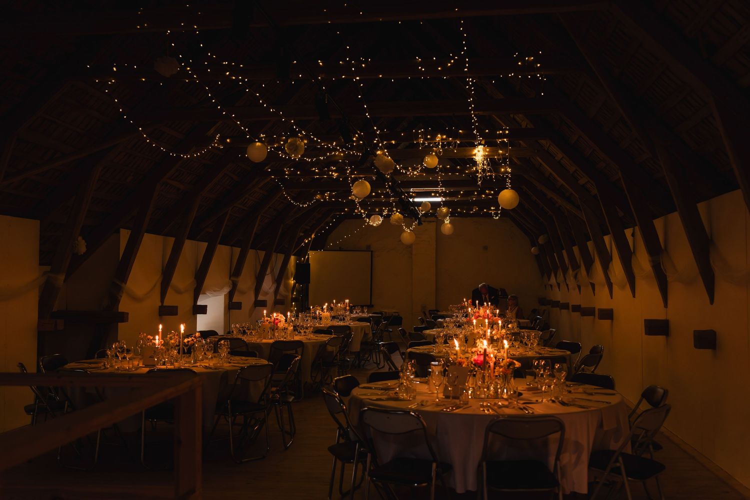 Rustic candlelit dining tables set up in a dimly lit barn venue with string lights creating a romantic atmosphere.