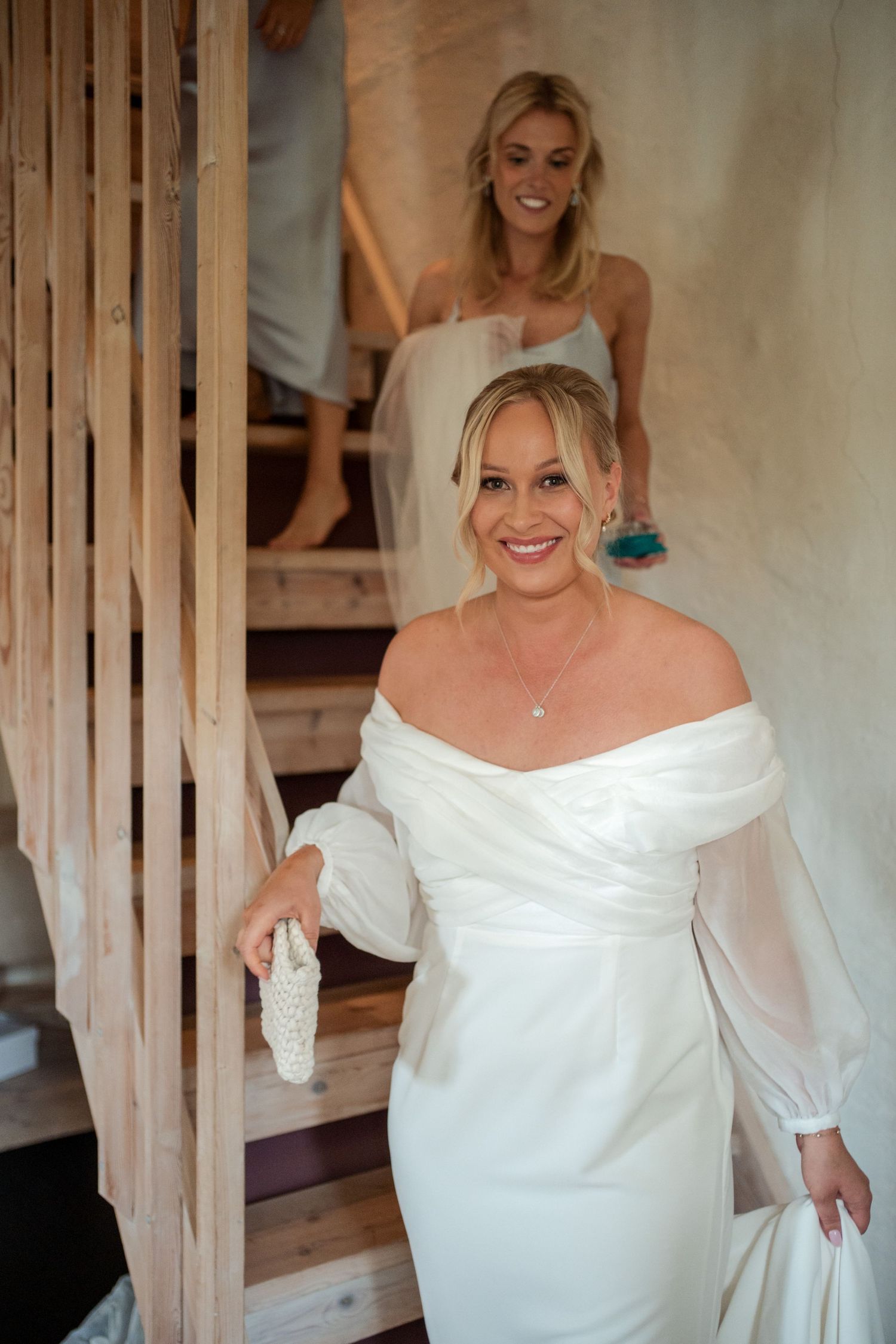 Bride in elegant off-shoulder white wedding dress with sheer sleeves descends wooden staircase while smiling.
