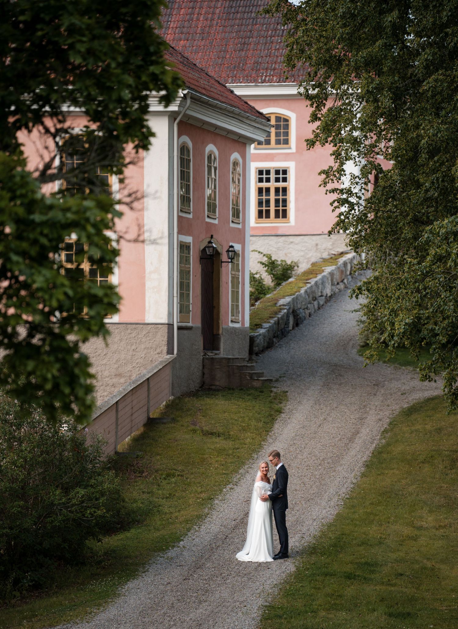 A couple stands on a gravel path beside a pink historic mansion with white trim and elegant architecture.