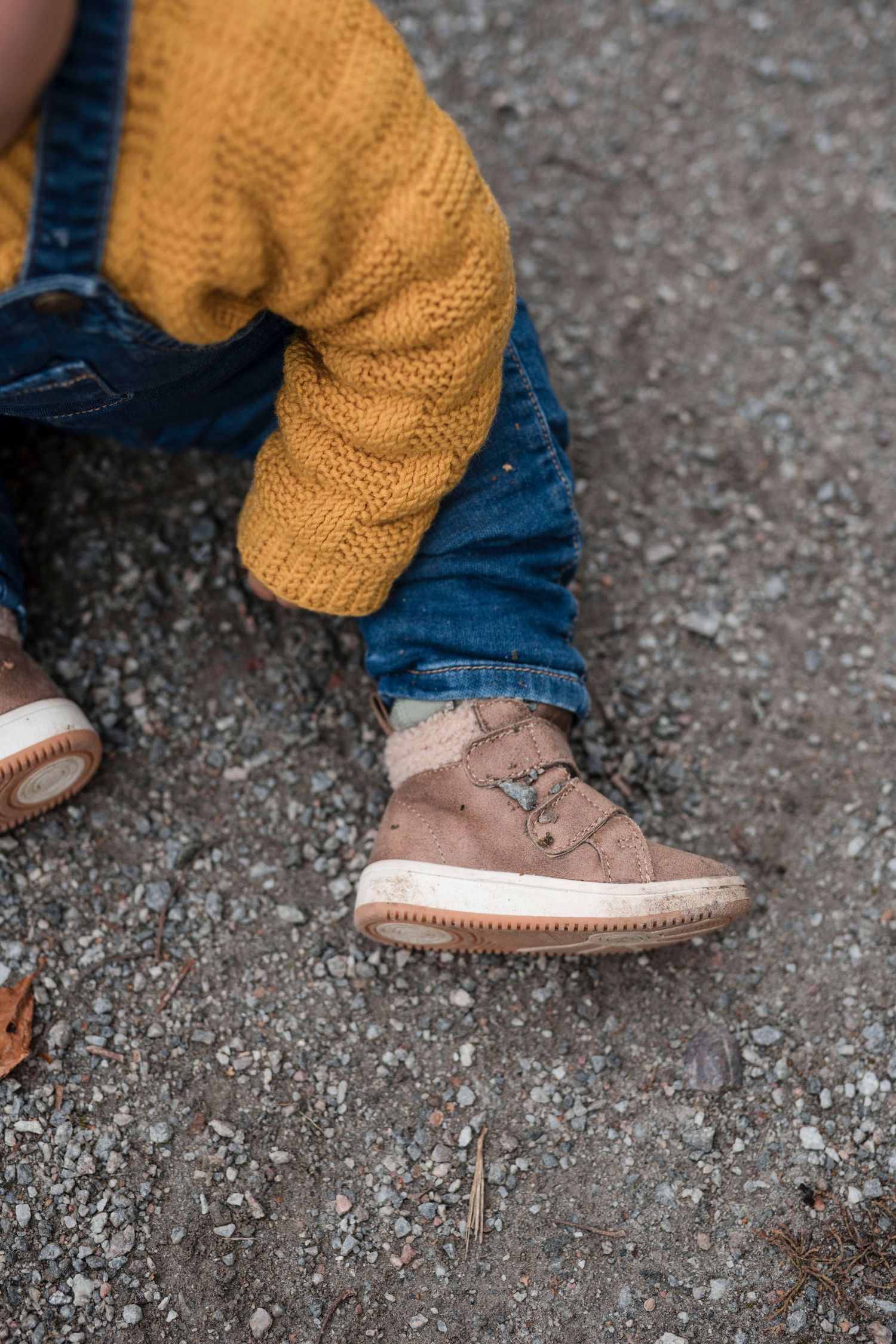 Close-up view of brown leather moccasin-style shoes paired with blue jeans and a mustard yellow sweater on gravel ground.