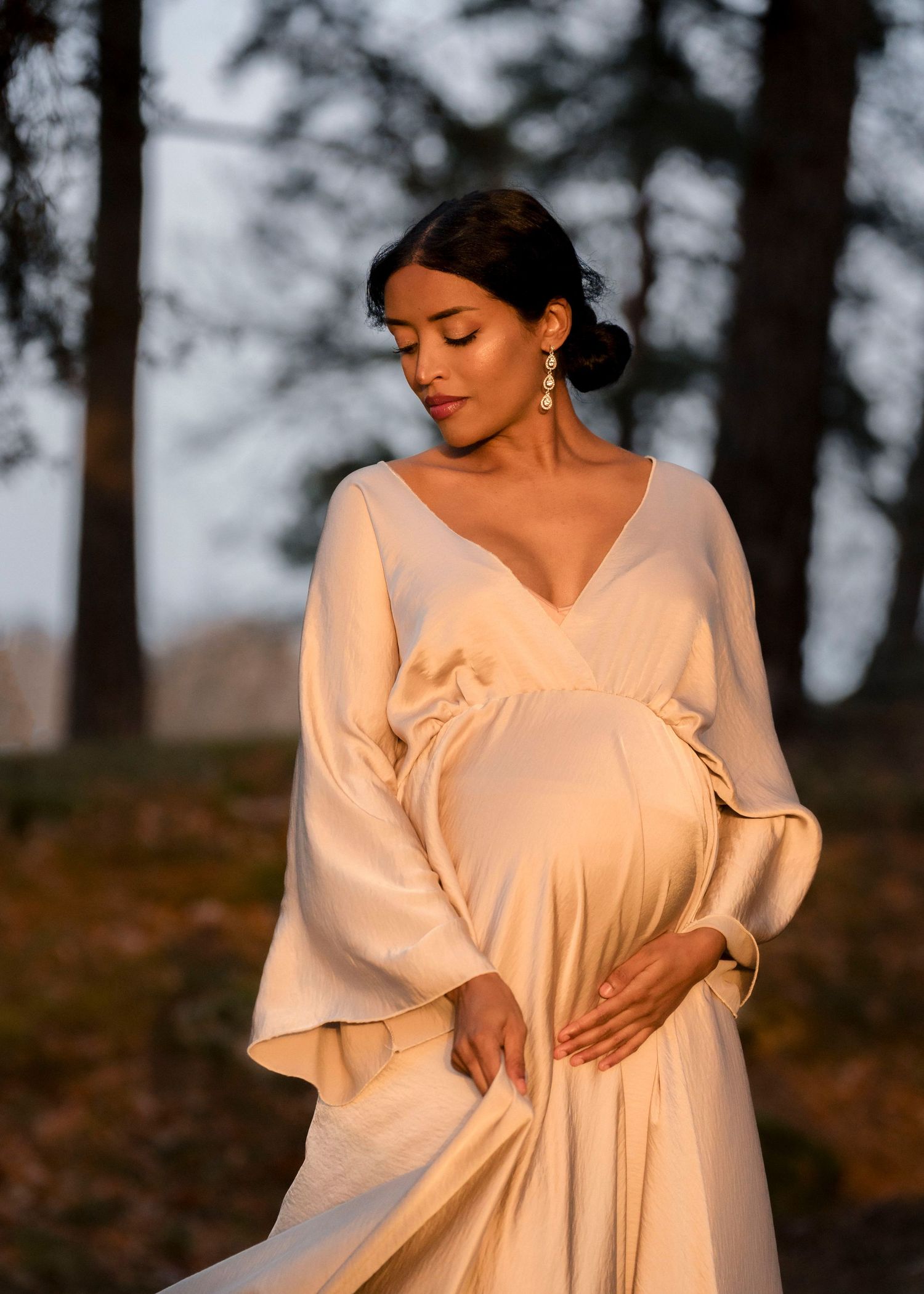 A person in a flowing cream-colored dress poses gracefully in a forest setting during golden hour.