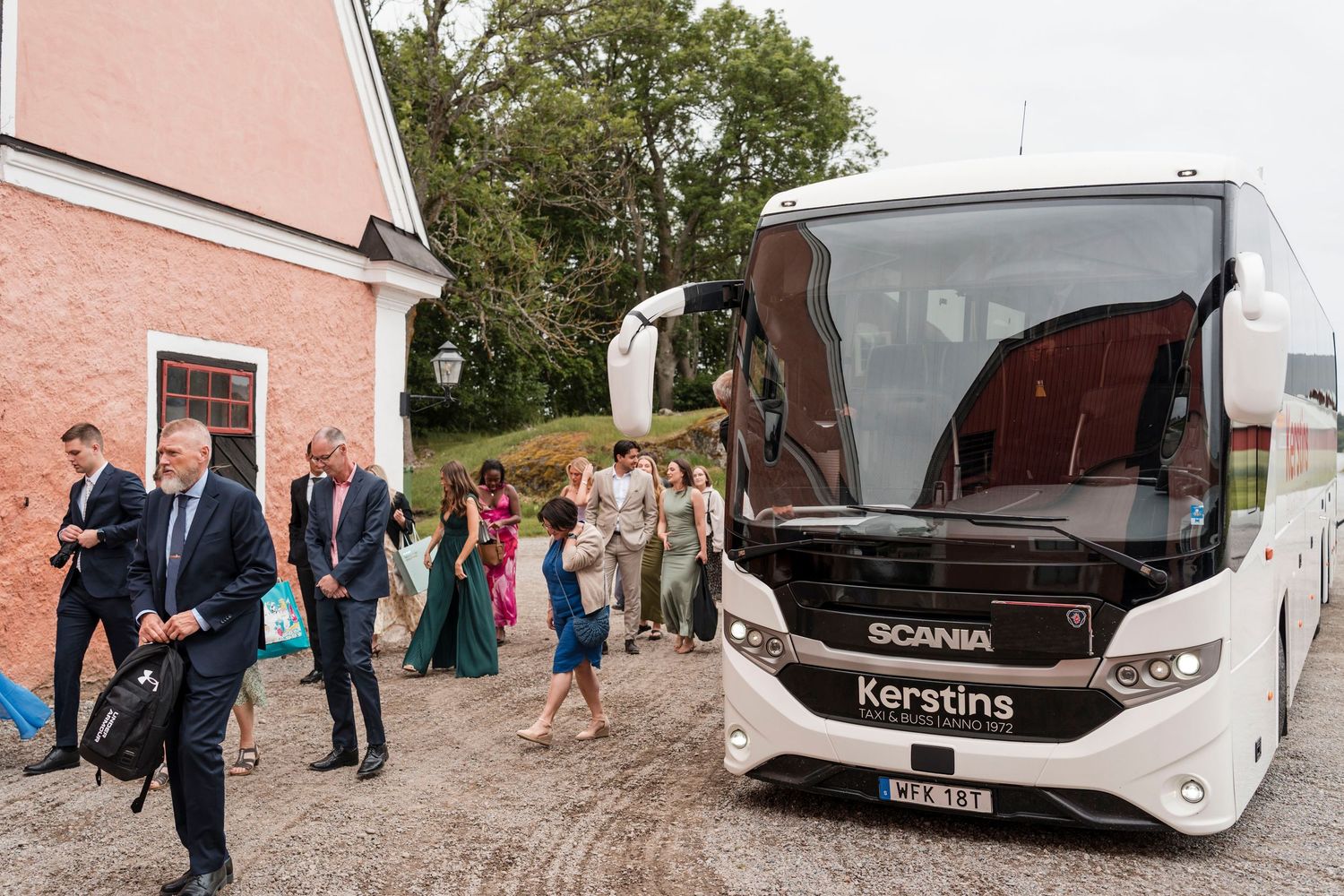 Wedding guests board a white charter bus parked outside a pink building on a sunny day.