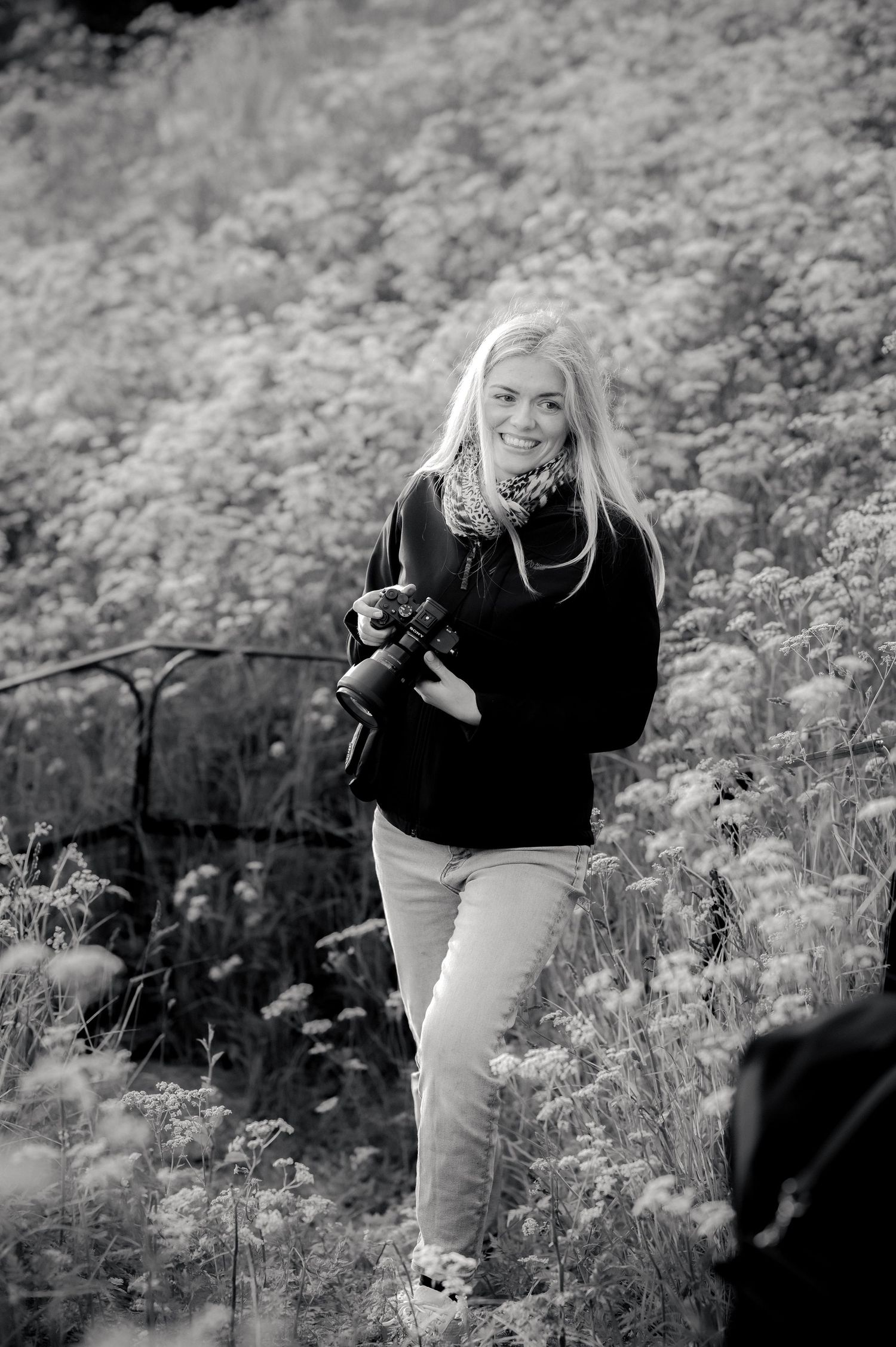 Black and white photo of photographer in black sweater and light pants holding camera while standing in nature.