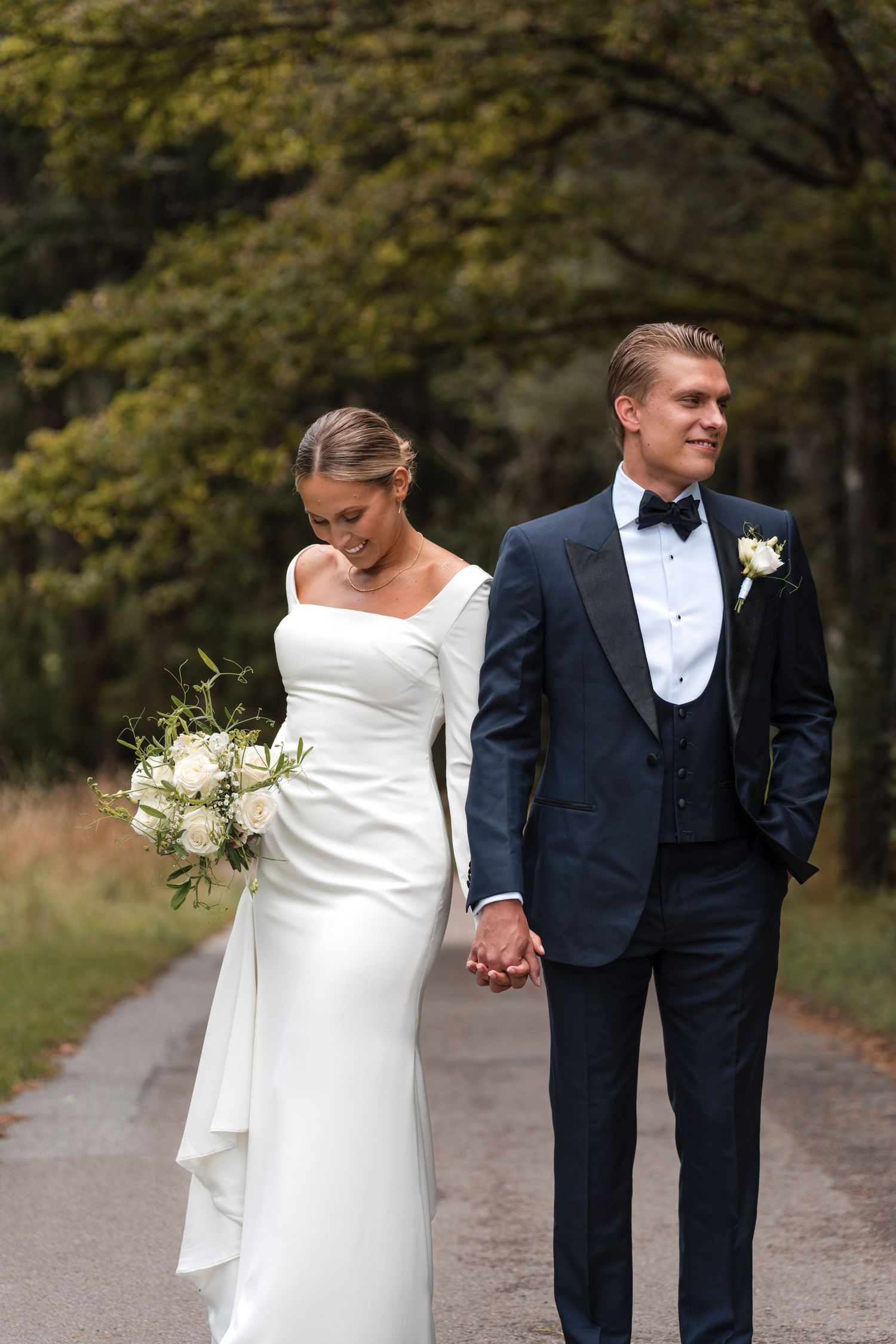 Newlywed couple walking hand in hand down a tree-lined autumn path in elegant wedding attire.