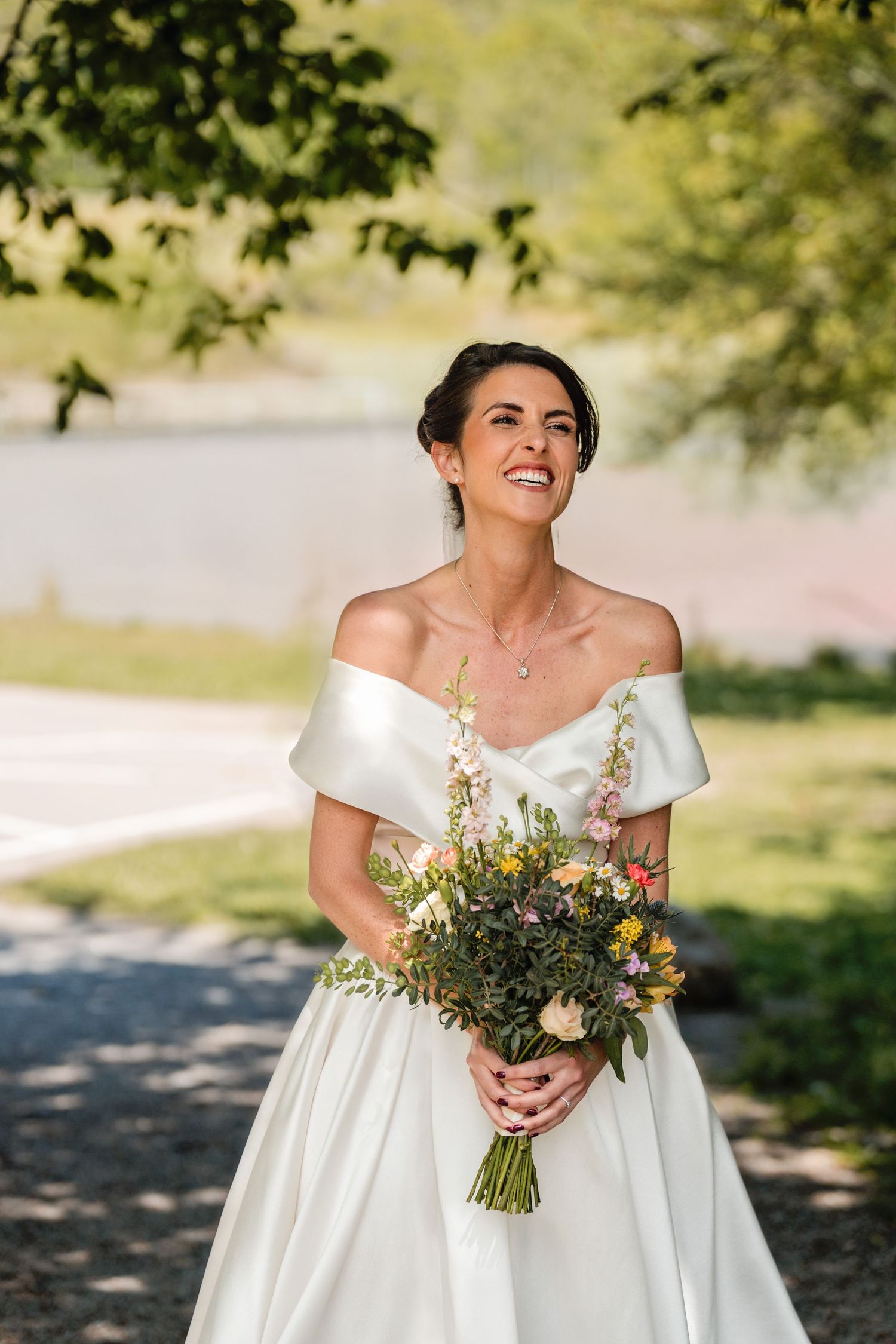 A bride in an off-shoulder white gown holds a colorful wildflower bouquet on a sunlit outdoor path.