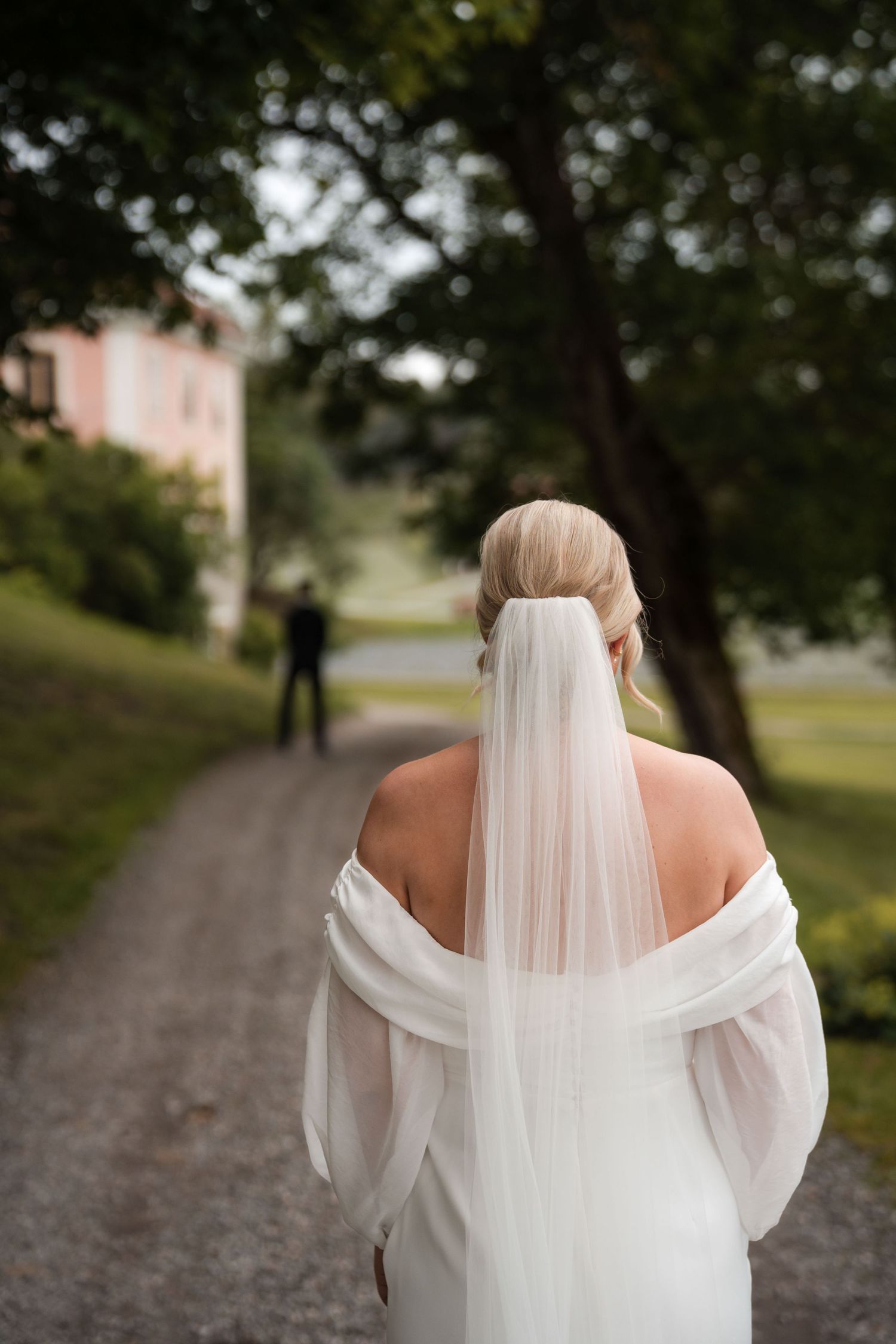 Back view of a bride in an elegant white off-shoulder wedding dress with a flowing veil walking along a tree-lined path.