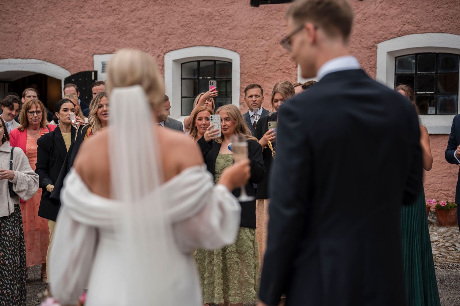 Wedding guests celebrate and take photos of the newlyweds during an outdoor reception at a brick venue.