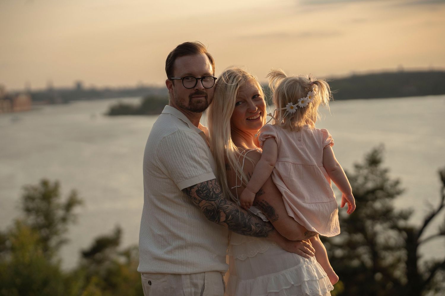 Couple in white outfits embracing on a beach at sunset with water and trees in the background.