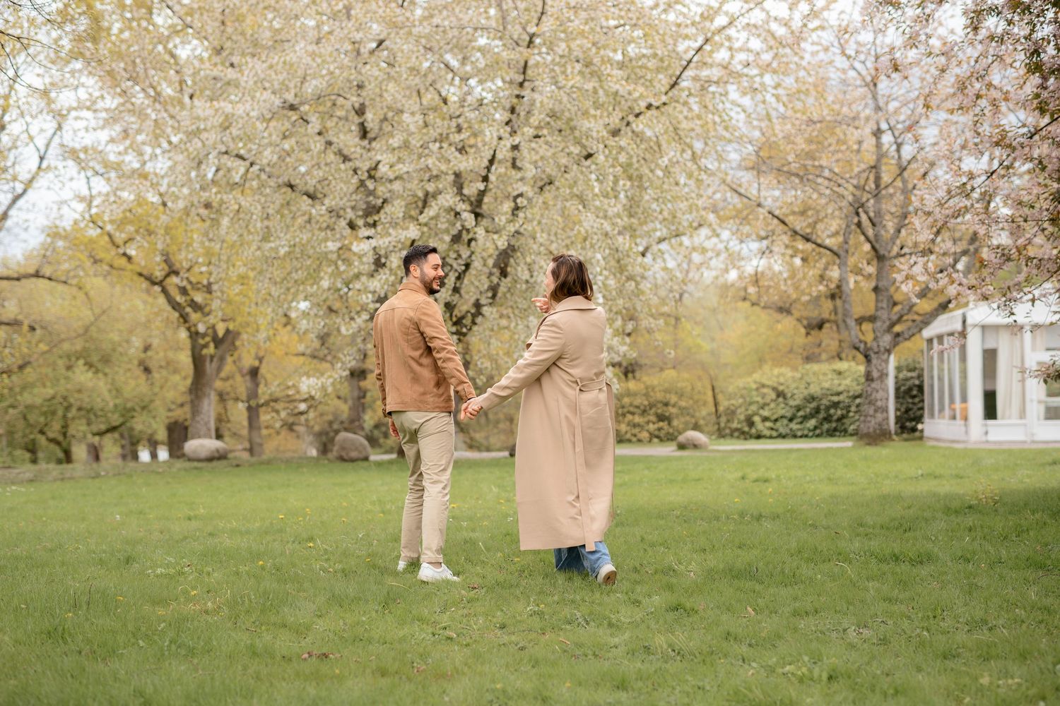 Two people hold hands while standing together on a green lawn surrounded by autumn trees.