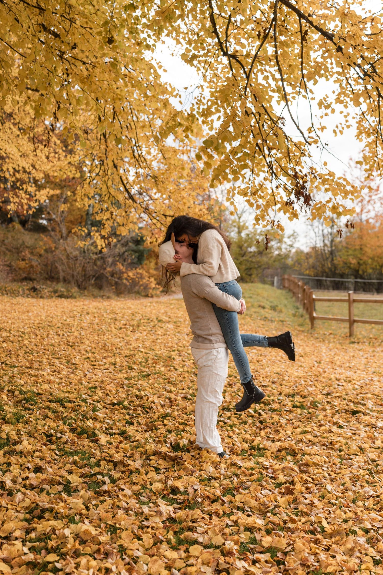 A couple embraces while standing in a carpet of golden autumn leaves on a fall afternoon in a park setting.